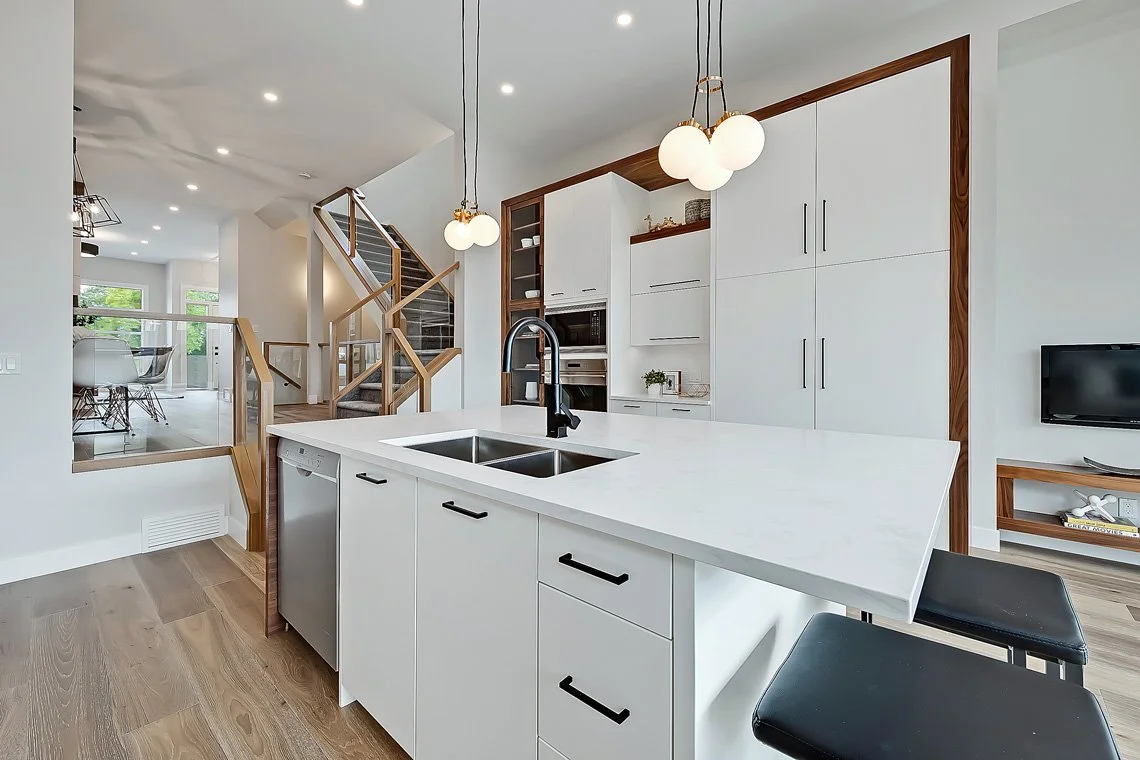 Modern kitchen with white cabinets, a white island with a sink and black handles, pendant lights hanging above, a built-in microwave, and a TV in the background.