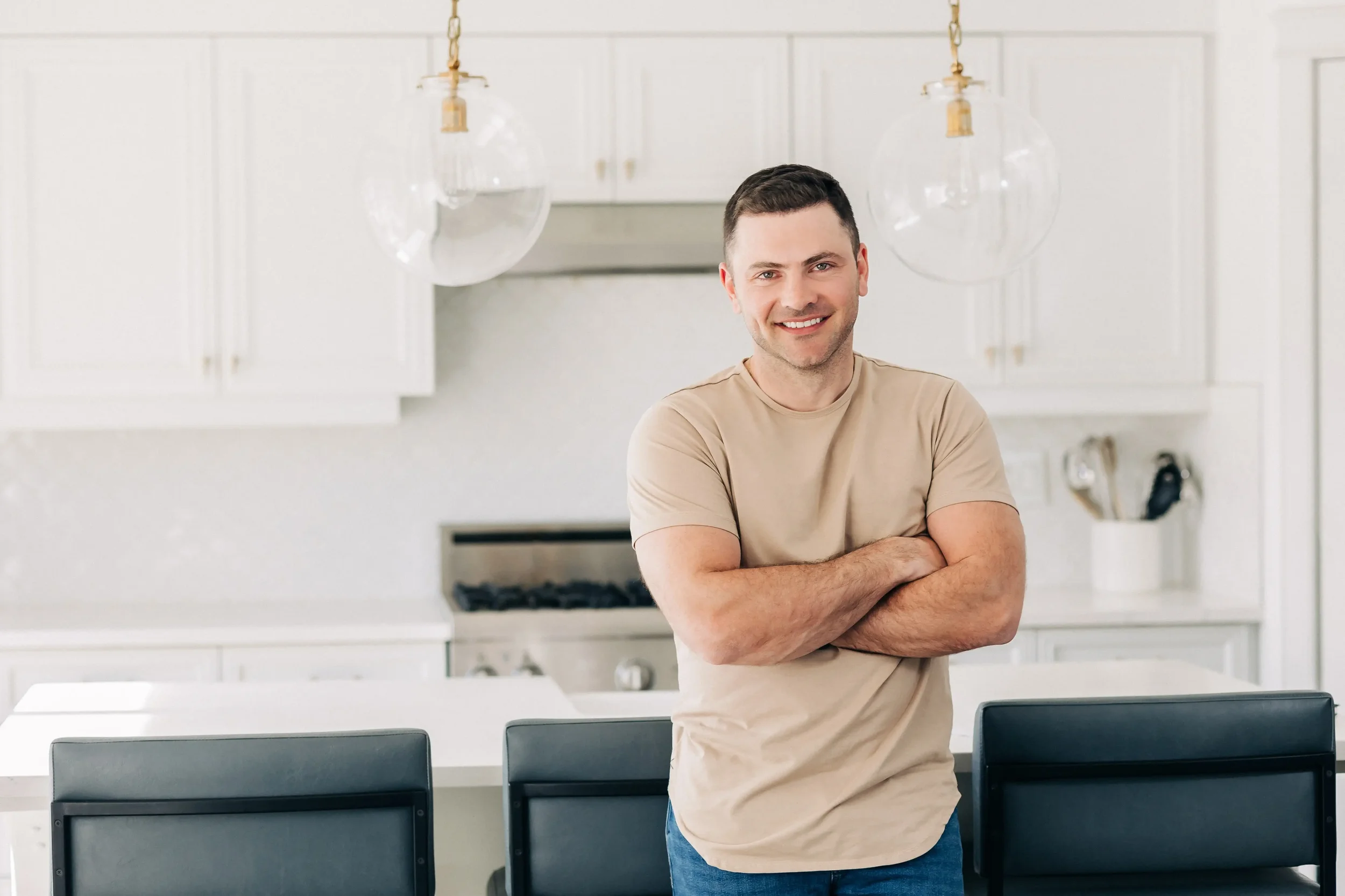 A young man with short dark hair and a light beard, smiling, standing with arms crossed in a bright, modern kitchen, wearing a beige t-shirt and blue jeans, with glass pendant lights hanging from the ceiling and white cabinetry in the background.
