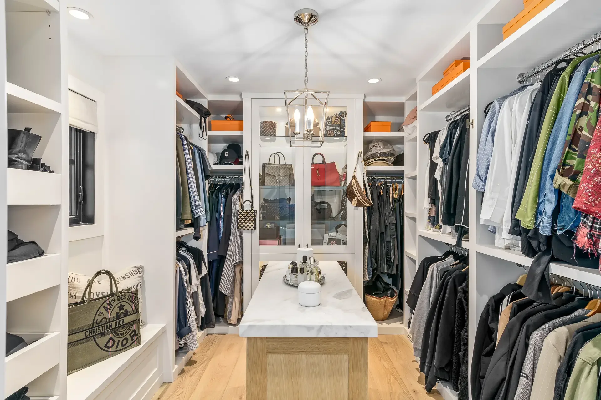 A walk-in closet with white built-in shelves filled with clothes, shoes, and boxes. In the center, there is a white marble-topped island with perfume bottles, a tissue box, and a small container. Overhead, there is a modern hanging light fixture with