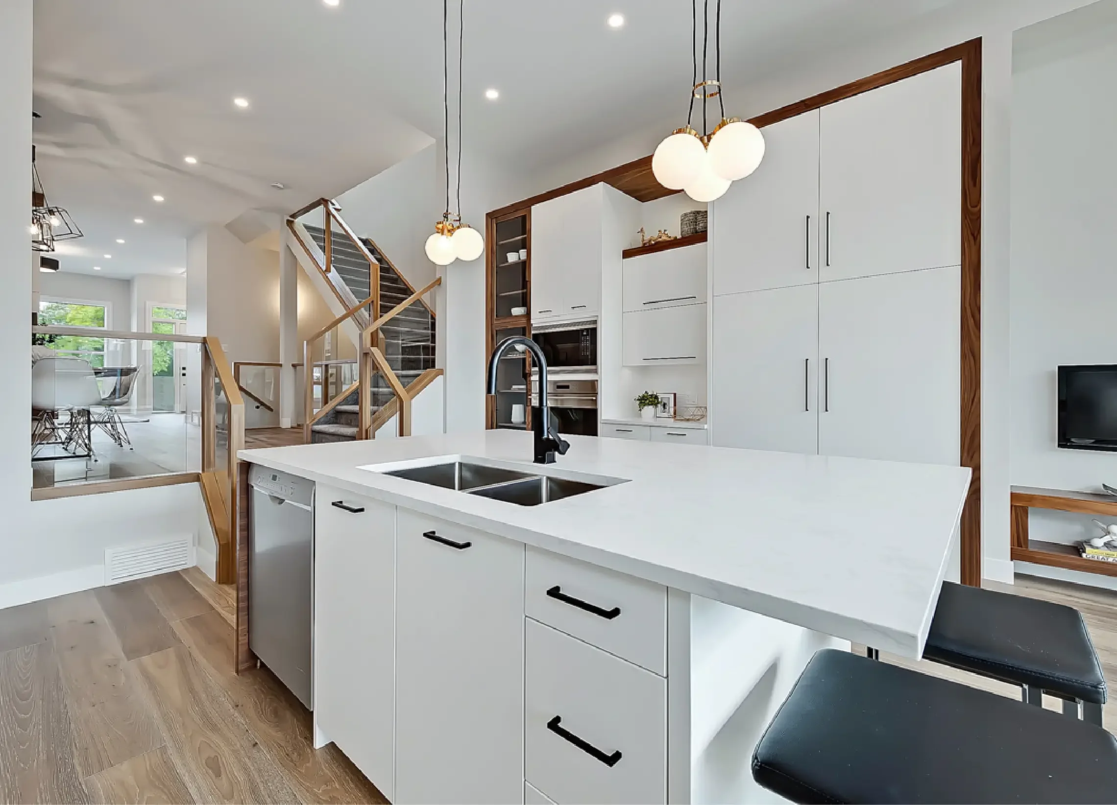 Modern kitchen with white cabinets, wooden accents, and a kitchen island with a black sink. There are two black chairs, a staircase with wooden railings, a dining area with glass table and white chairs, and large windows letting in natural light.