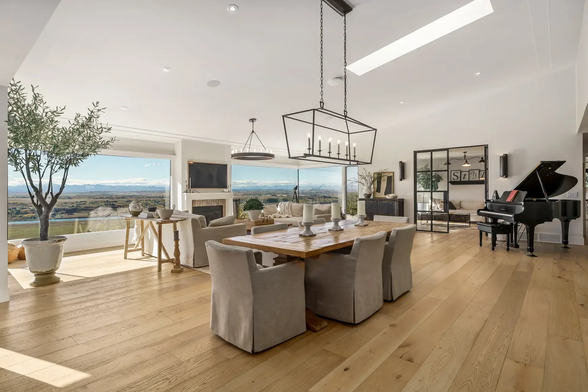 Open-concept living and dining room with large windows showcasing mountain and lake views, light wood flooring, a black grand piano, and modern decor.