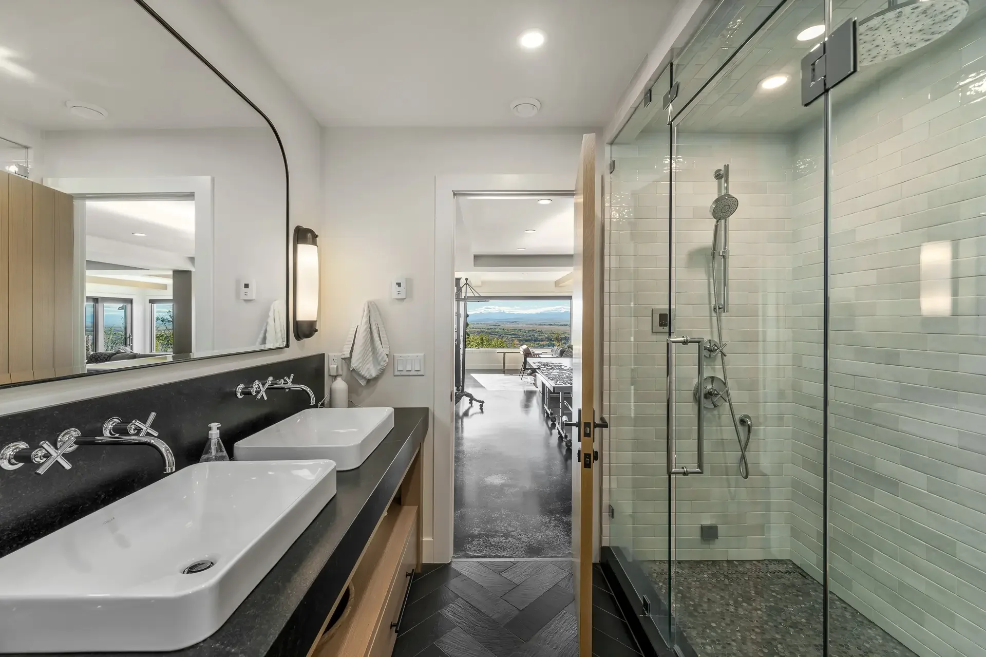 Modern bathroom with dual sinks, large mirror, and walk-in shower with glass door, overlooking a room with large windows and a landscape view.