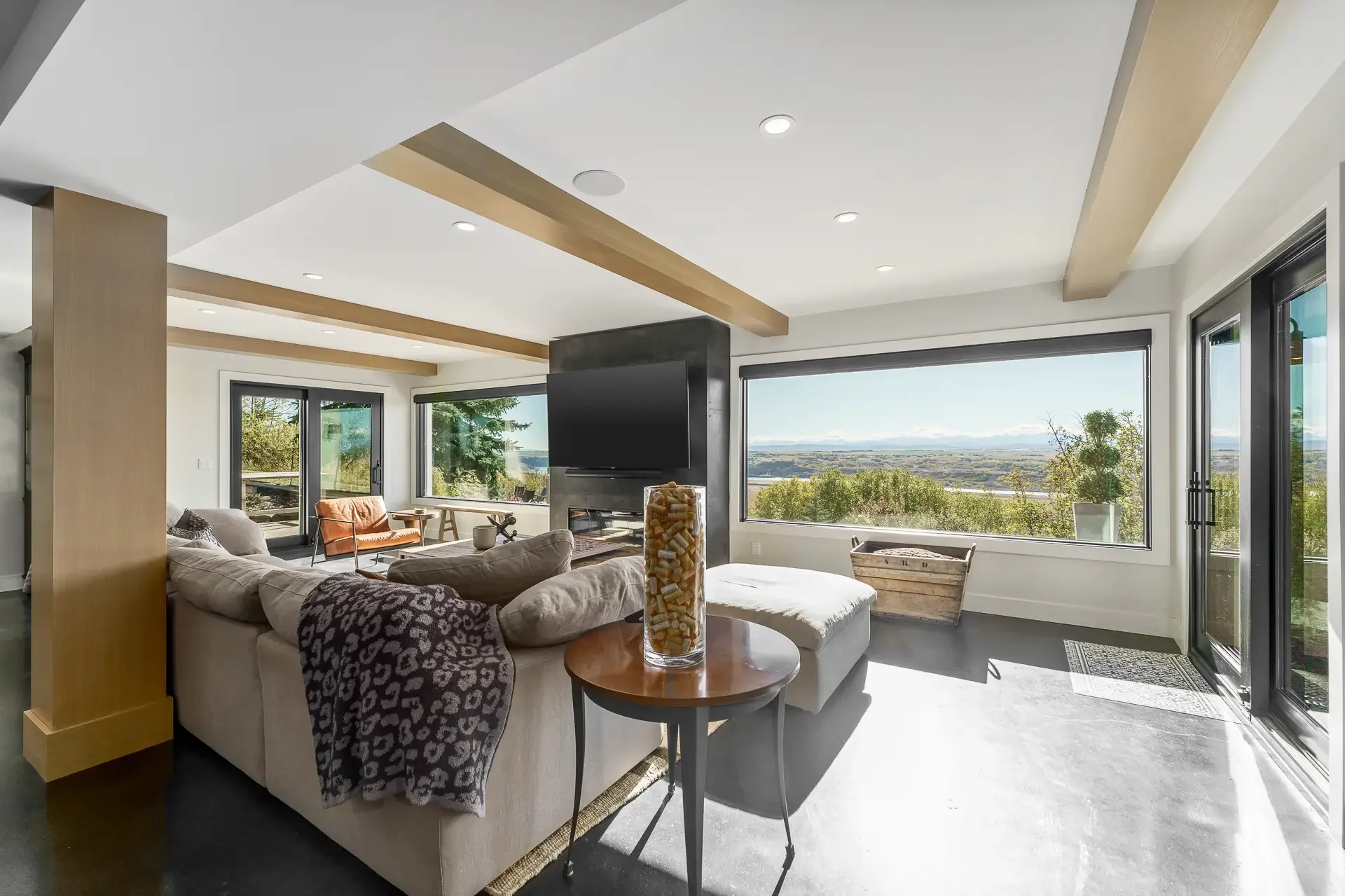 Living room with large windows showing a scenic view of trees and sky, white sofa with patterned throw, orange chair, TV, and decorative items.