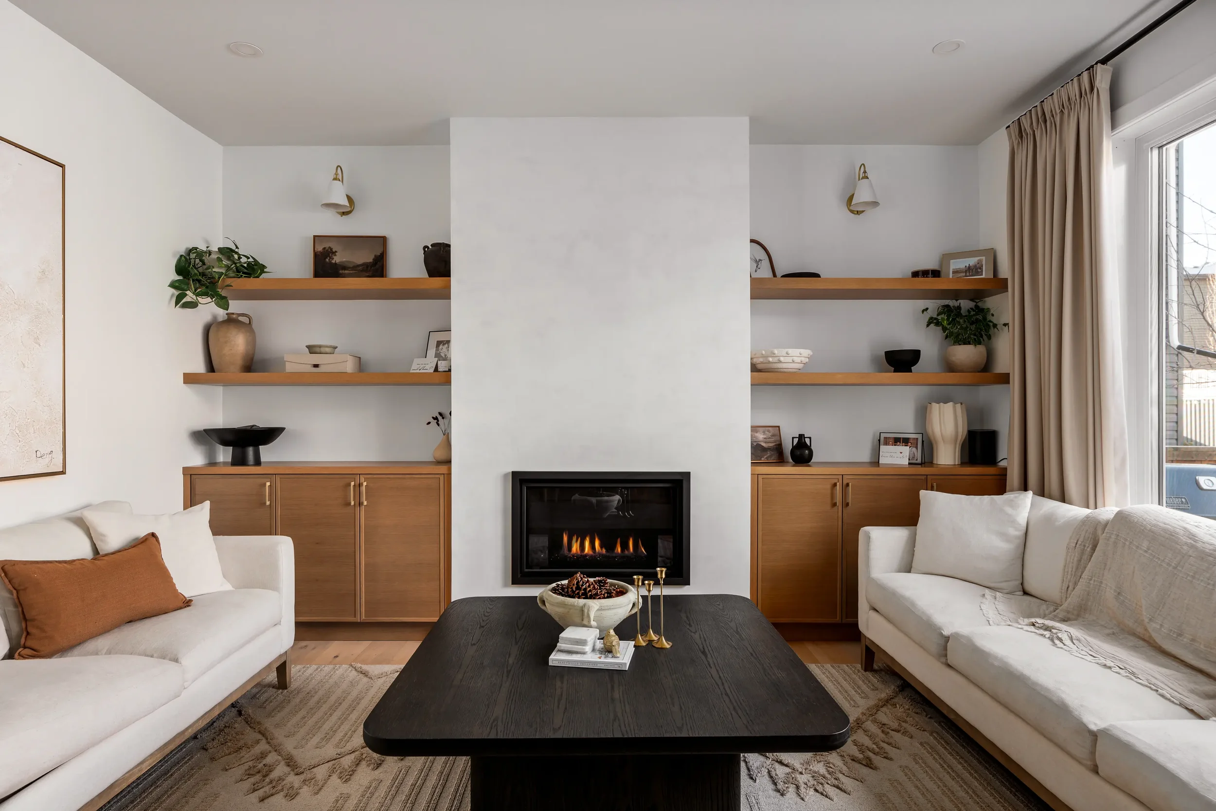 Living room with a fireplace, two white sofas, a black coffee table, built-in shelves with plants and decor, and a window with curtains.