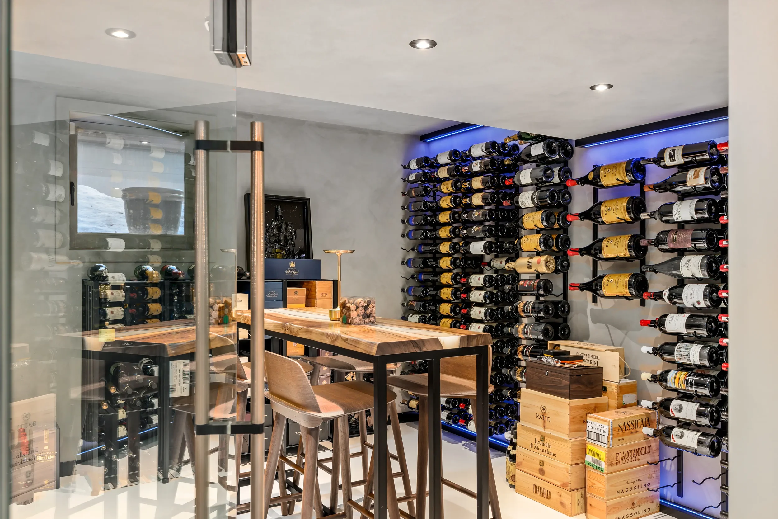 Wine cellar with wine bottles stored on black racks, a wooden table with chairs, and wine boxes on the floor, illuminated by ceiling lights and blue LED strip lighting.