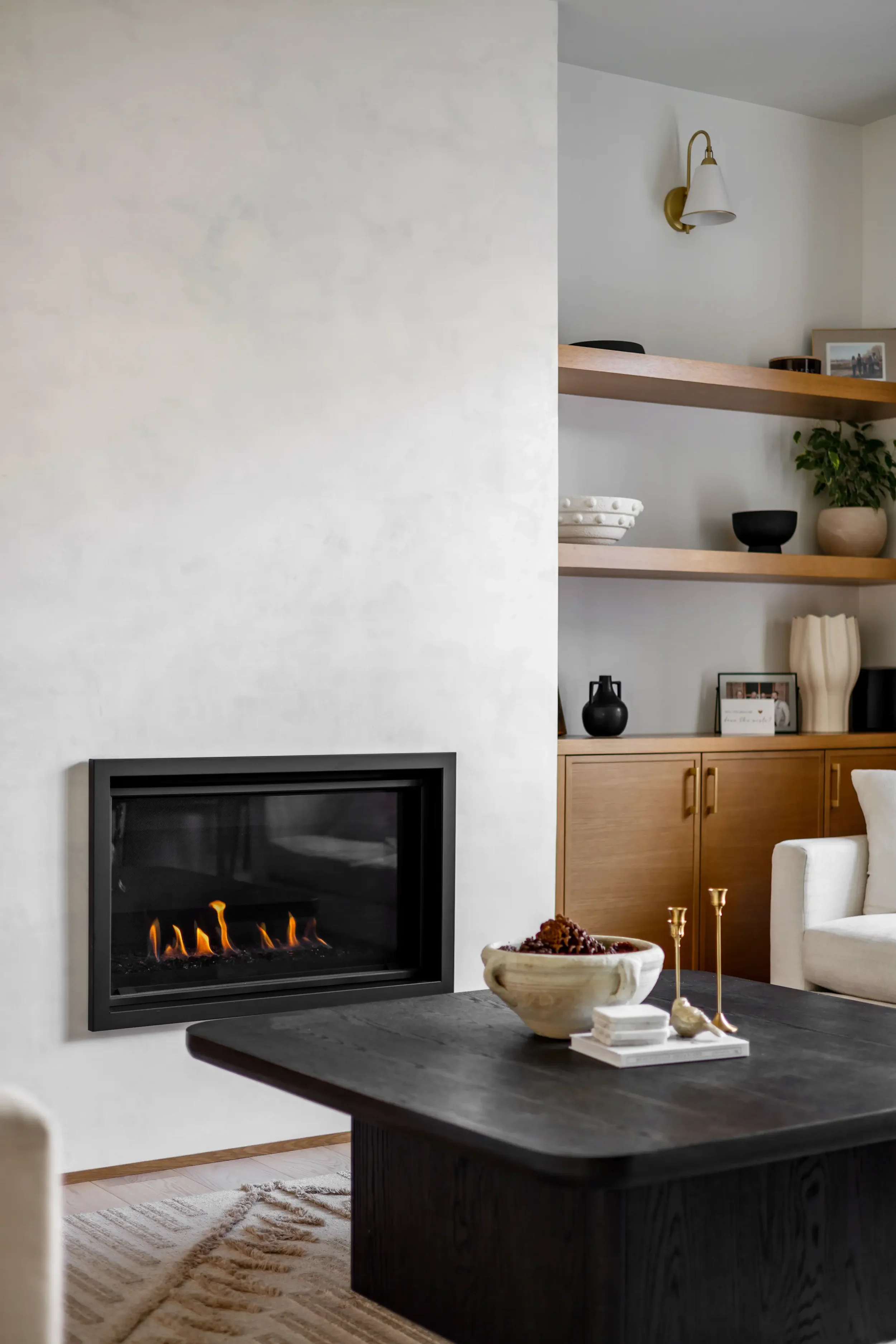 Living room with a fireplace, a dark wooden table with decorative items, a white rug, built-in wooden shelves with decor and framed photos, and a beige armchair.