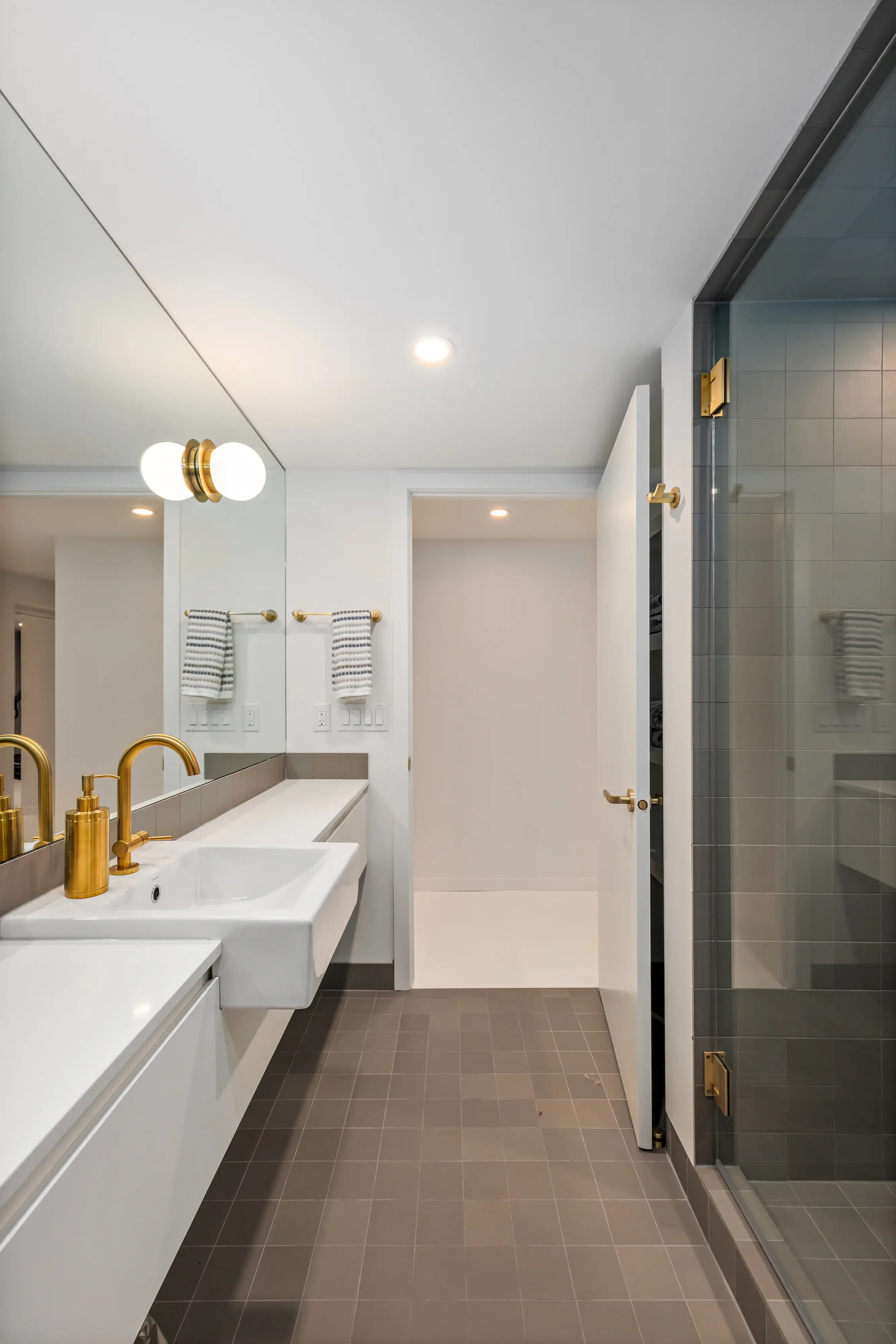 Modern bathroom with white walls, gray floor tiles, a white sink with gold fixtures, large mirror, two white and gray striped towels on a gold towel bar, ceiling lights, and a glass shower enclosure with gray tiles.