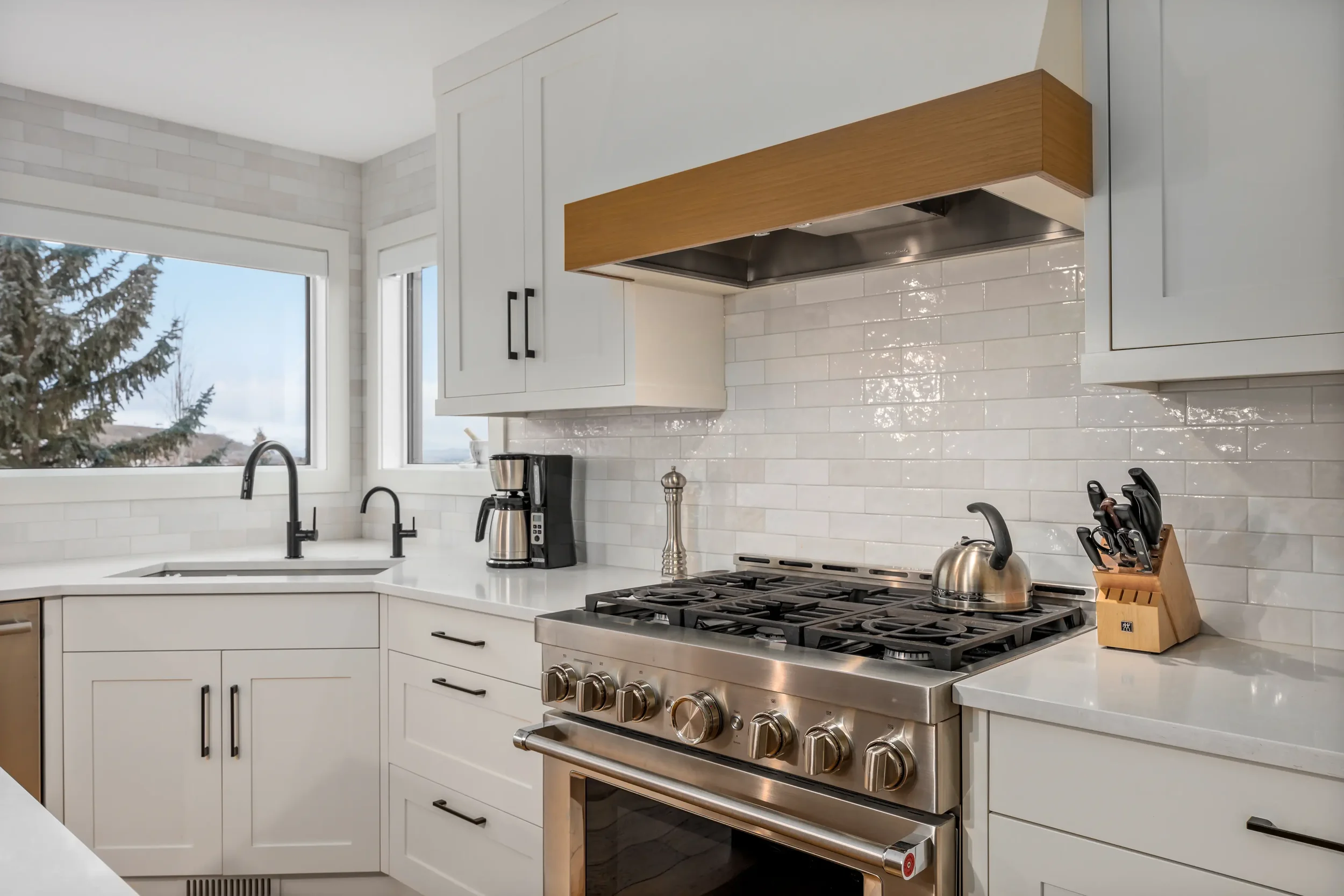 Modern white kitchen with stainless steel gas stove, black faucet, white cabinets, and a window overlooking trees.