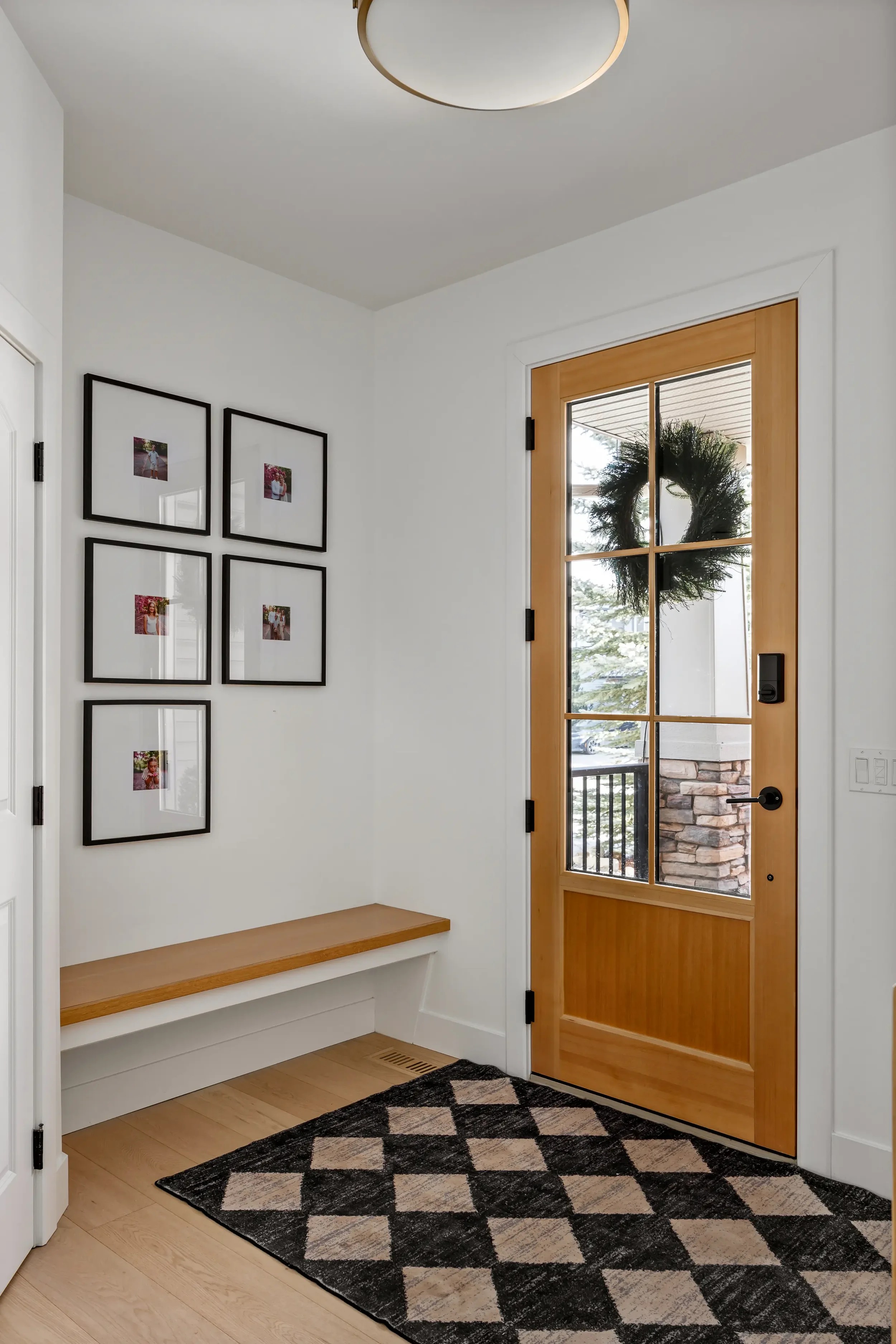 Entryway with a wooden door featuring glass panels and a wreath. White walls with black-framed photos, a wooden bench, a black and beige patterned rug, and a ceiling light.