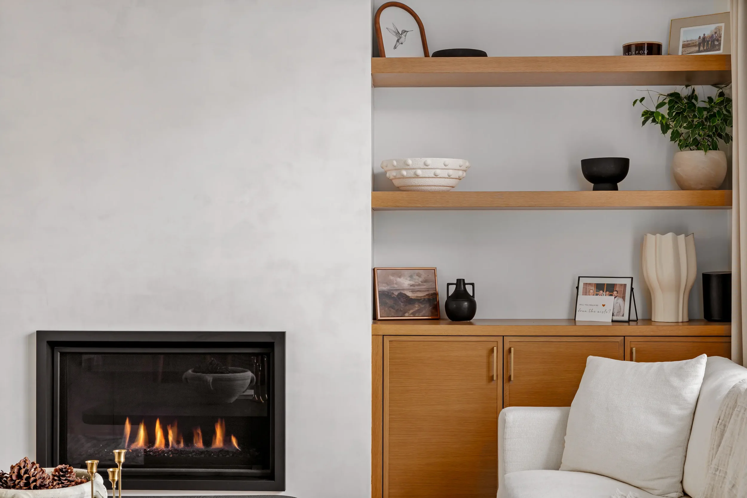 Living room with modern fireplace, wooden bookshelf with decorative items, framed photos, a plant, and a white sofa with pillows.