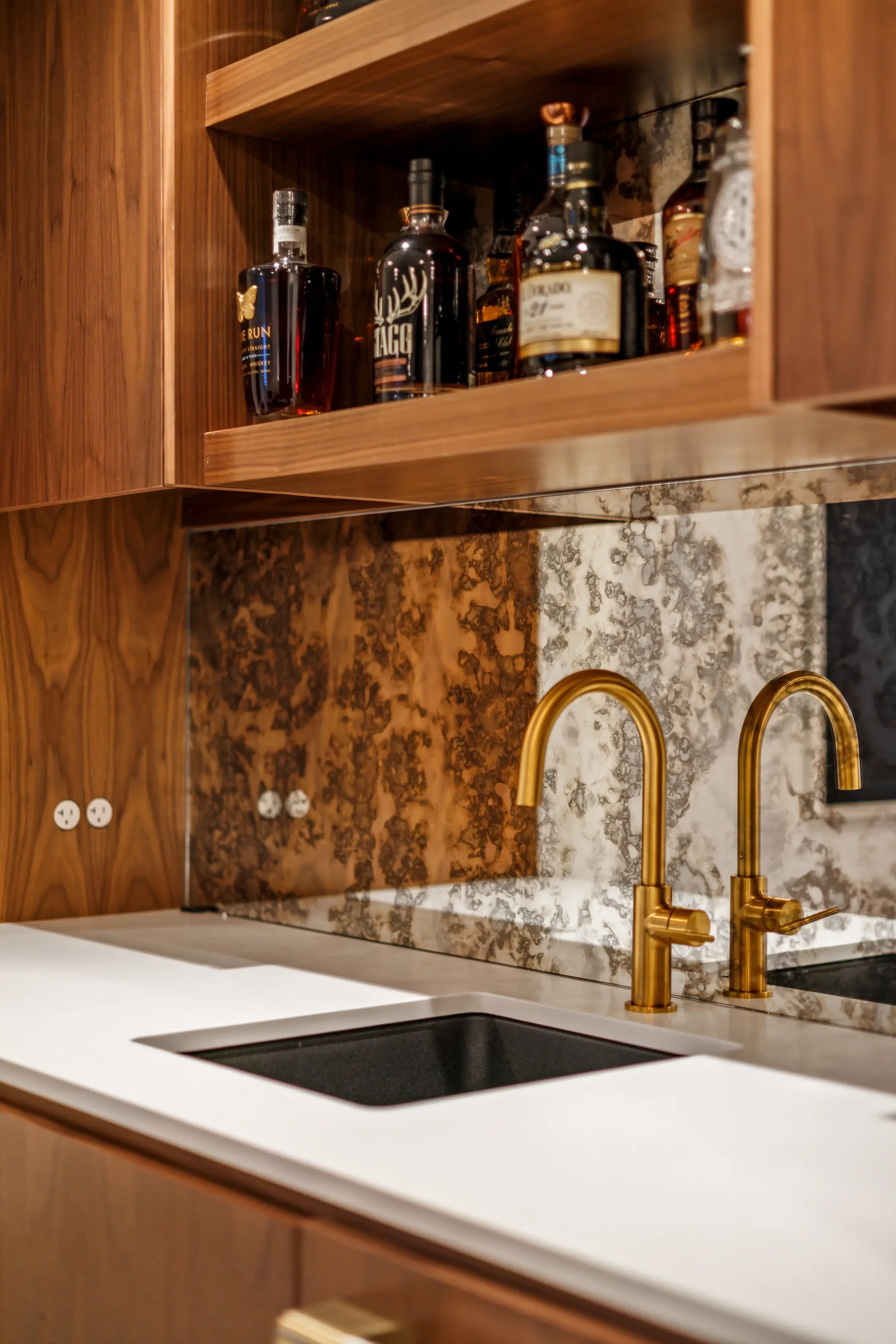 Close-up of a kitchen countertop with a black sink, gold faucet, marble backsplash with gold accents, and wooden cabinets.