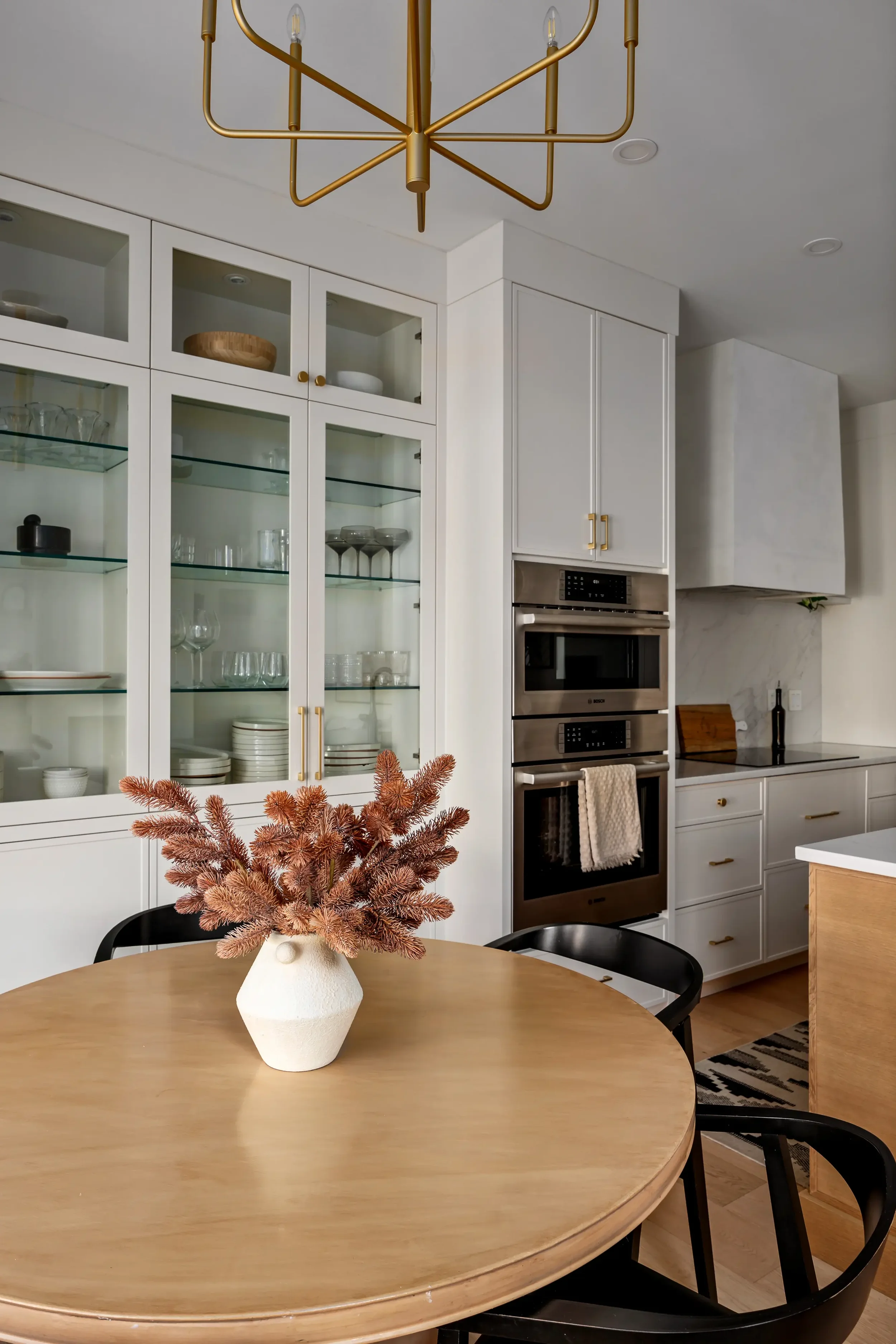 A white kitchen with glass-front cabinets displaying glassware, a gold chandelier hanging from the ceiling, a wooden dining table with a white vase holding pinkish-brown berry branches, and black chairs around the table.