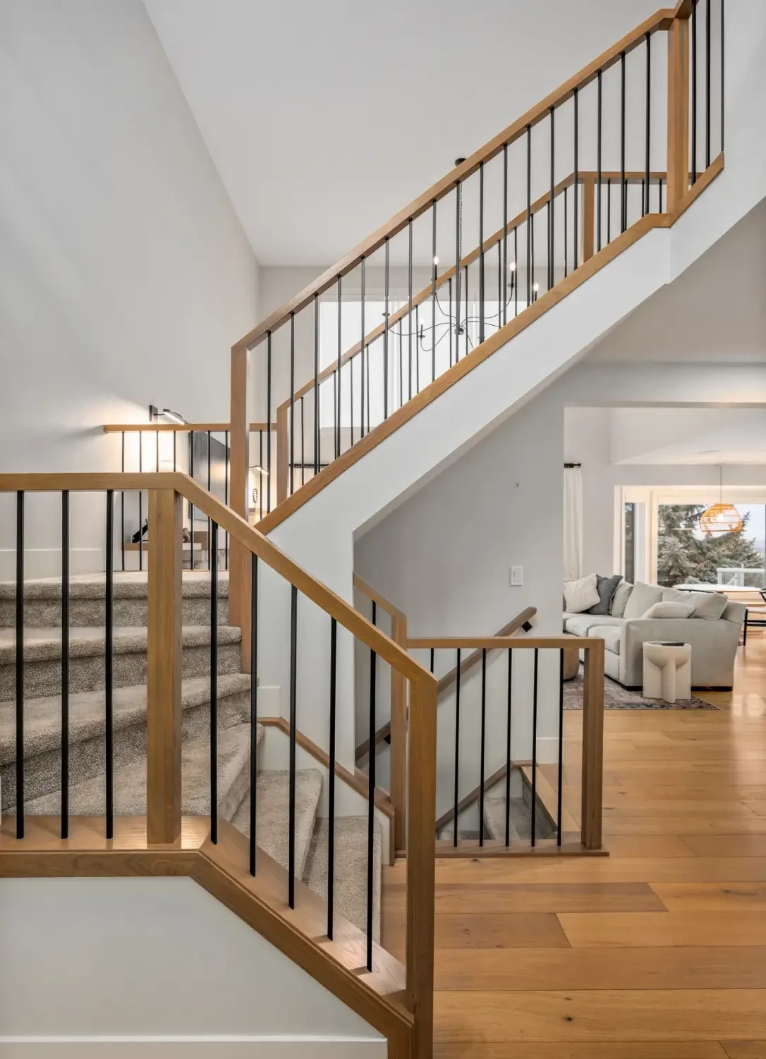 Interior of a modern home featuring a staircase with wooden handrails and black balusters, leading to an upper floor, with a living room visible in the background including a sofa and a large window.