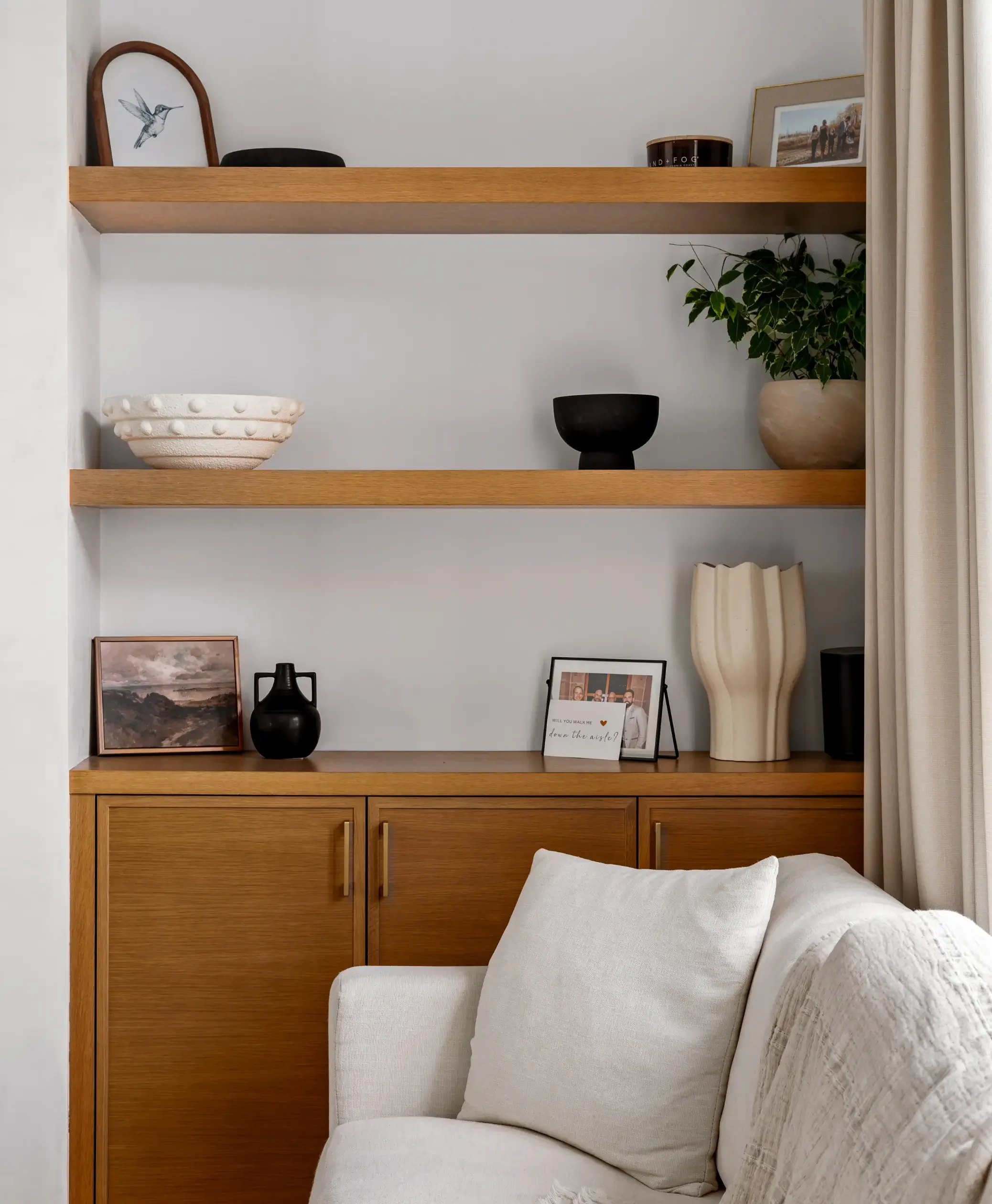 A living room with wooden wall shelves holding decorative items, framed photos, a potted plant, a black vase, and a framed landscape picture. There is a beige curtain on the right and a white sofa with plush pillows in the foreground.
