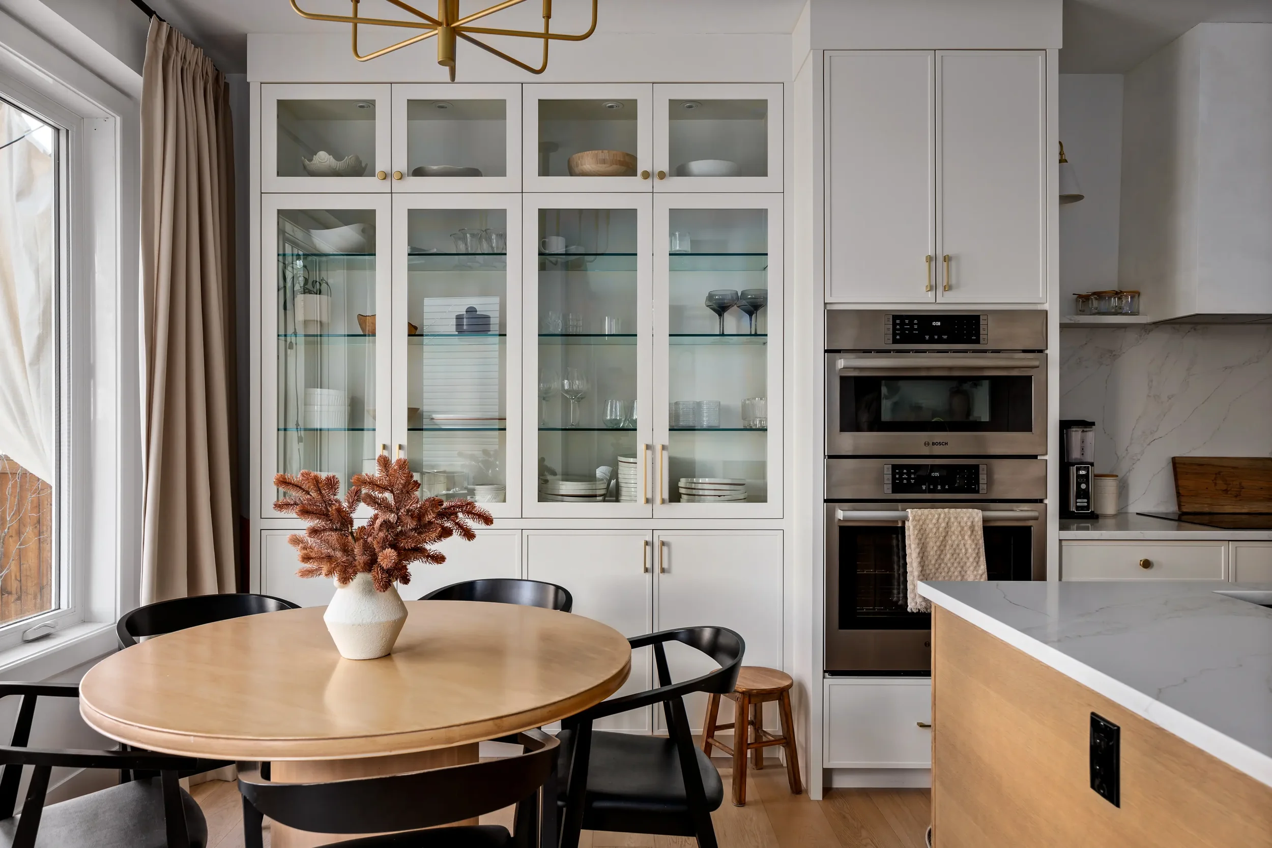 A modern kitchen with white cabinetry, glass-front upper cabinets, stainless steel appliances, a round wooden dining table with black chairs, and a vase with pink dried flowers on the table. There's a window with beige curtains on the left.