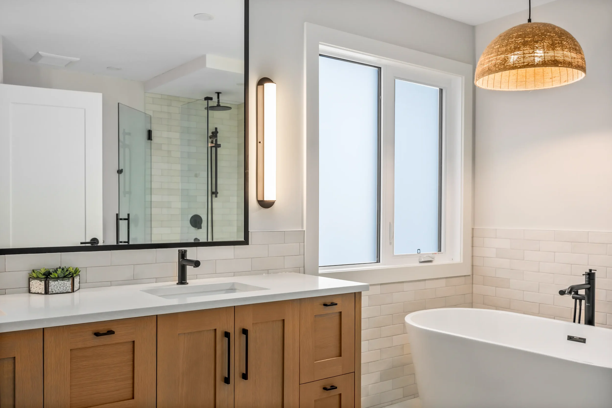 Modern bathroom with a large mirror, double vanity with wooden cabinets, white countertop, black fixtures, and a rectangular window. Features a walk-in shower with glass doors and a hanging woven light fixture.