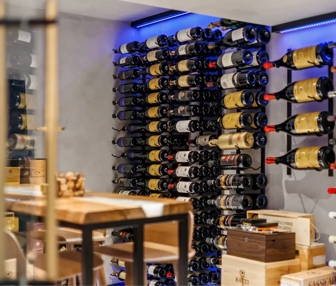 Wine bottles stored horizontally on a wall-mounted rack in a wine cellar or shop, with some wooden wine crates and shelves visible in the foreground.