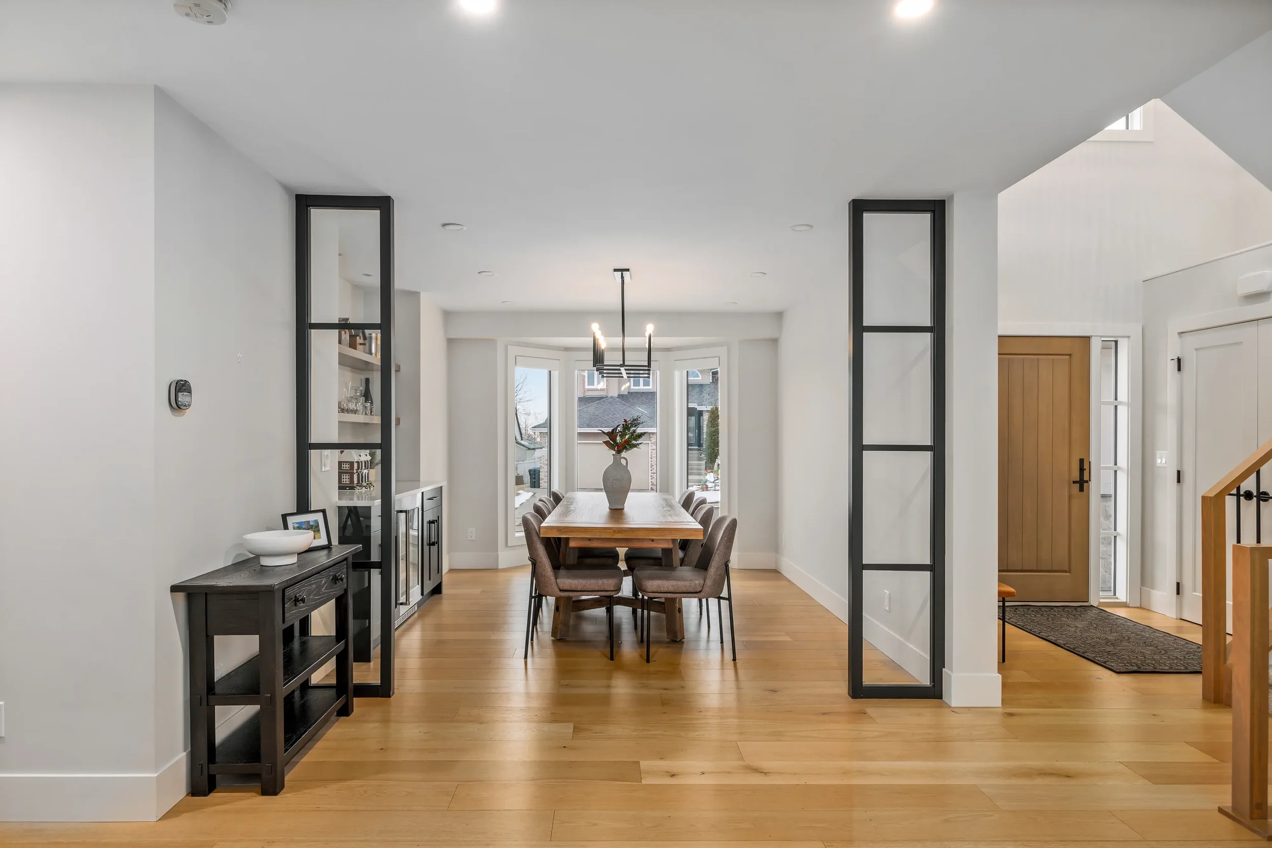 Open-concept living and dining area with white walls, wooden flooring, a long wooden dining table with chairs, a black sideboard, a vase with a plant, a window at the back, and modern light fixtures.
