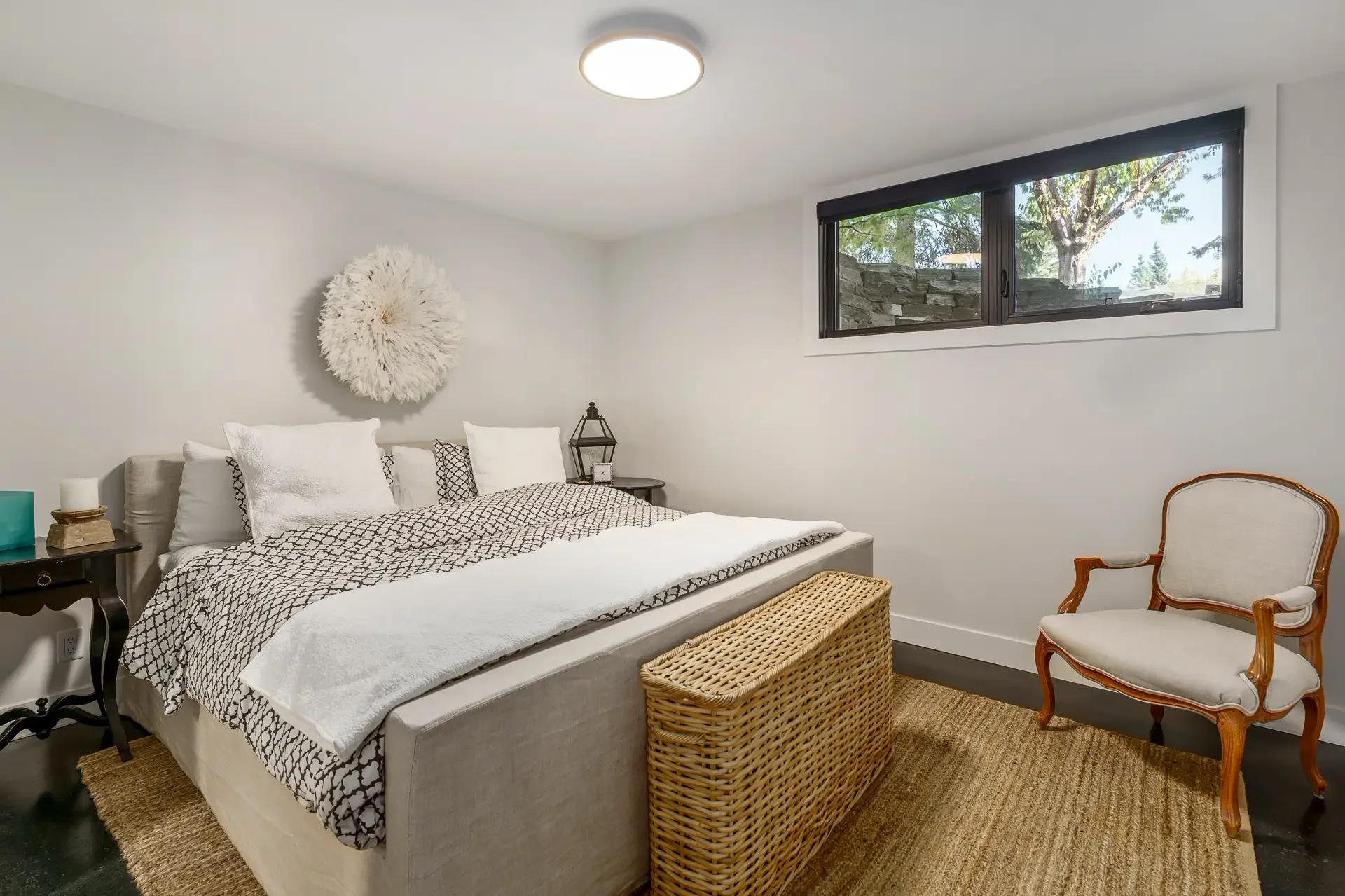 A modern bedroom with a bed, nightstand, and a decorative wall hanging. The room has a window with a view of trees outside, and an armchair with wooden arms and a cushion.