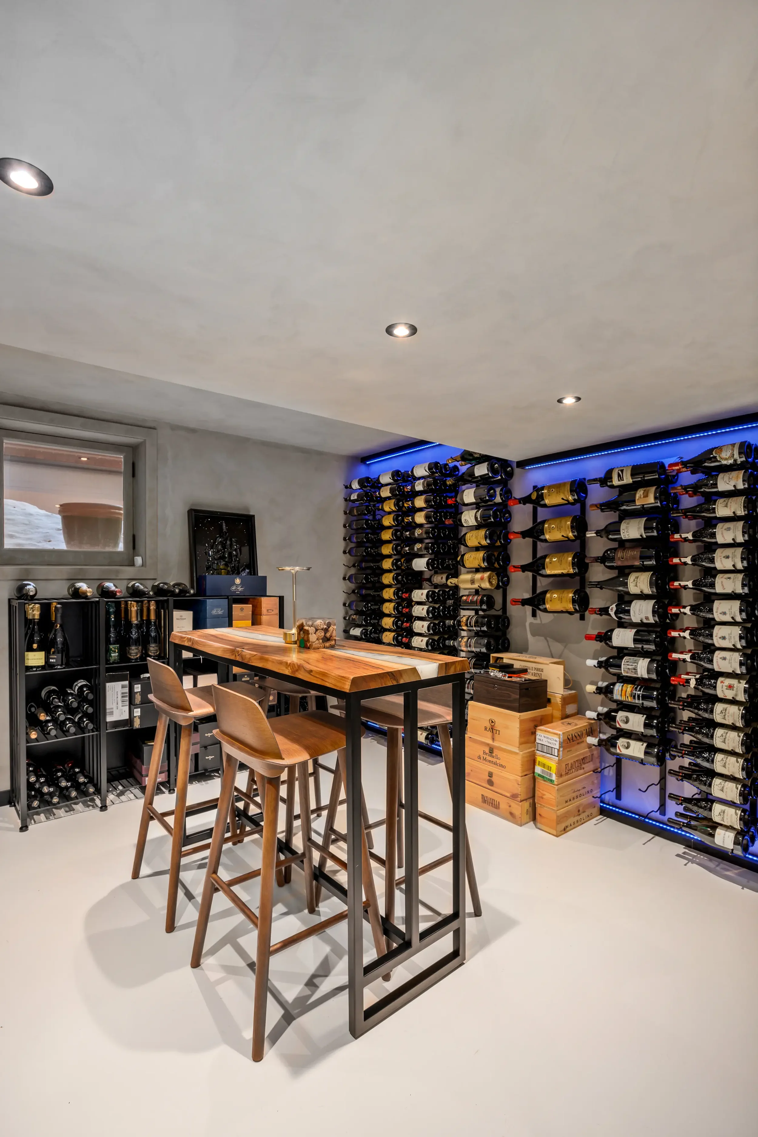 A cozy wine cellar with bottles stored on wall-mounted racks, a wooden table with chairs, a small window, and wine crates on the floor, illuminated by ceiling lights and ambient blue LED lighting.