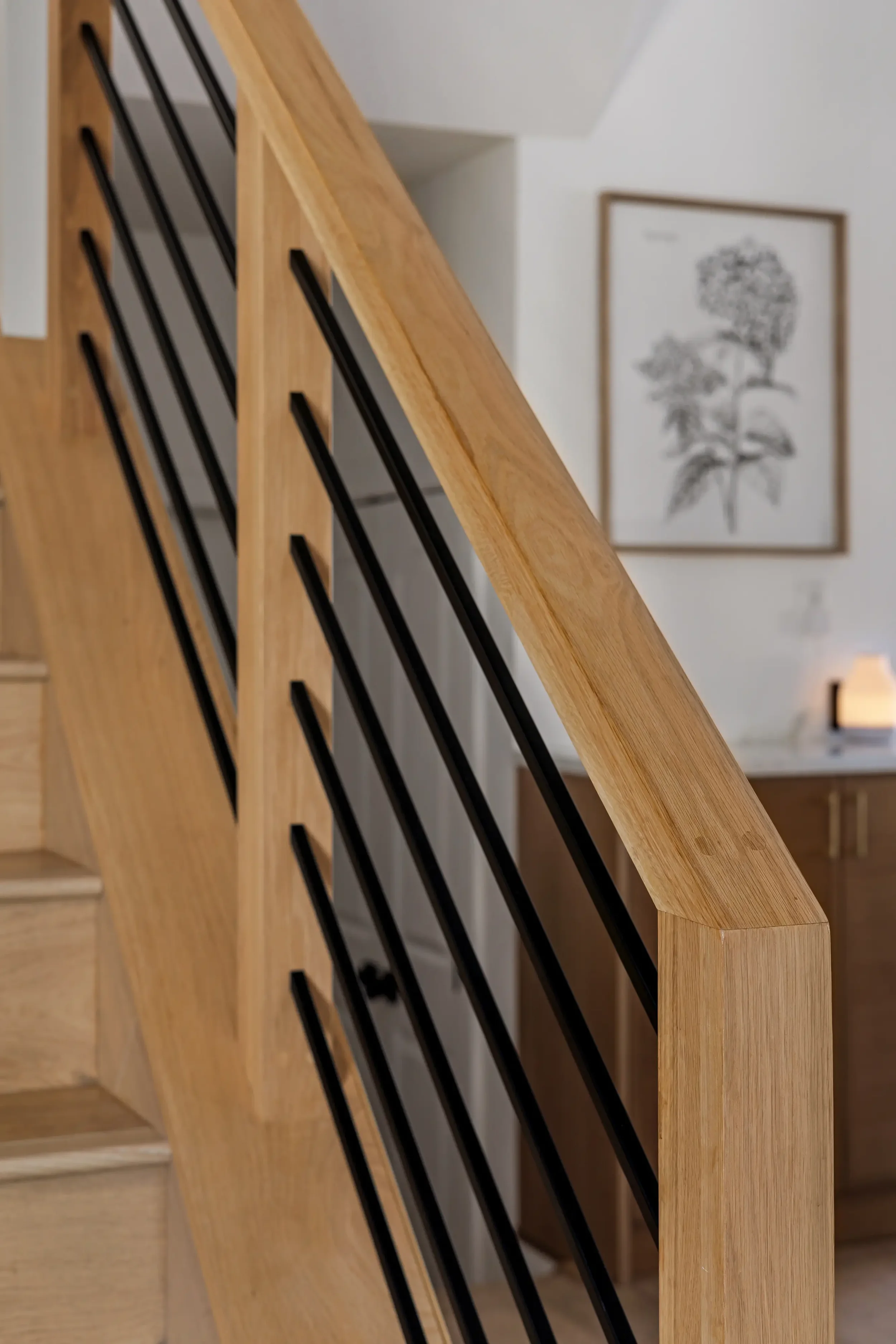 Close-up of a wooden staircase with black metal railings, with a framed botanical illustration on a white wall in the background.