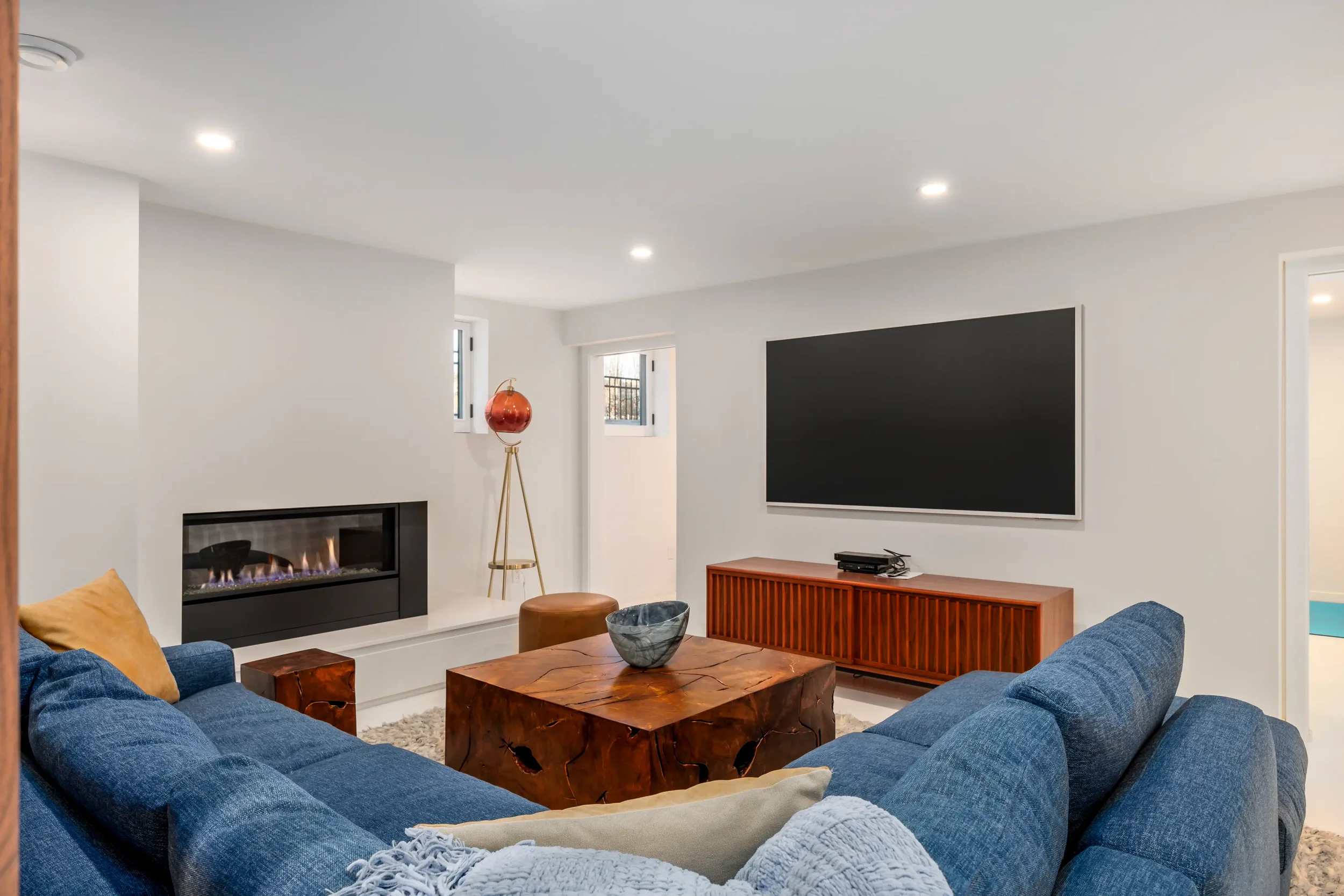 Living room with modern decor including a blue sectional sofa, wooden coffee table, wall-mounted flat-screen TV, fireplace, window with natural light, and a red decorative floor lamp.