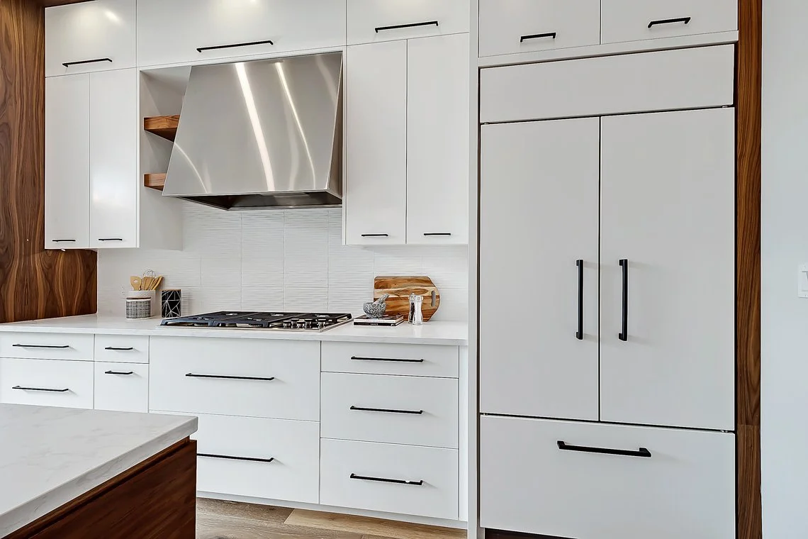 Modern kitchen with white cabinets, stainless steel range hood, and a built-in refrigerator, featuring black handles and wooden accents.