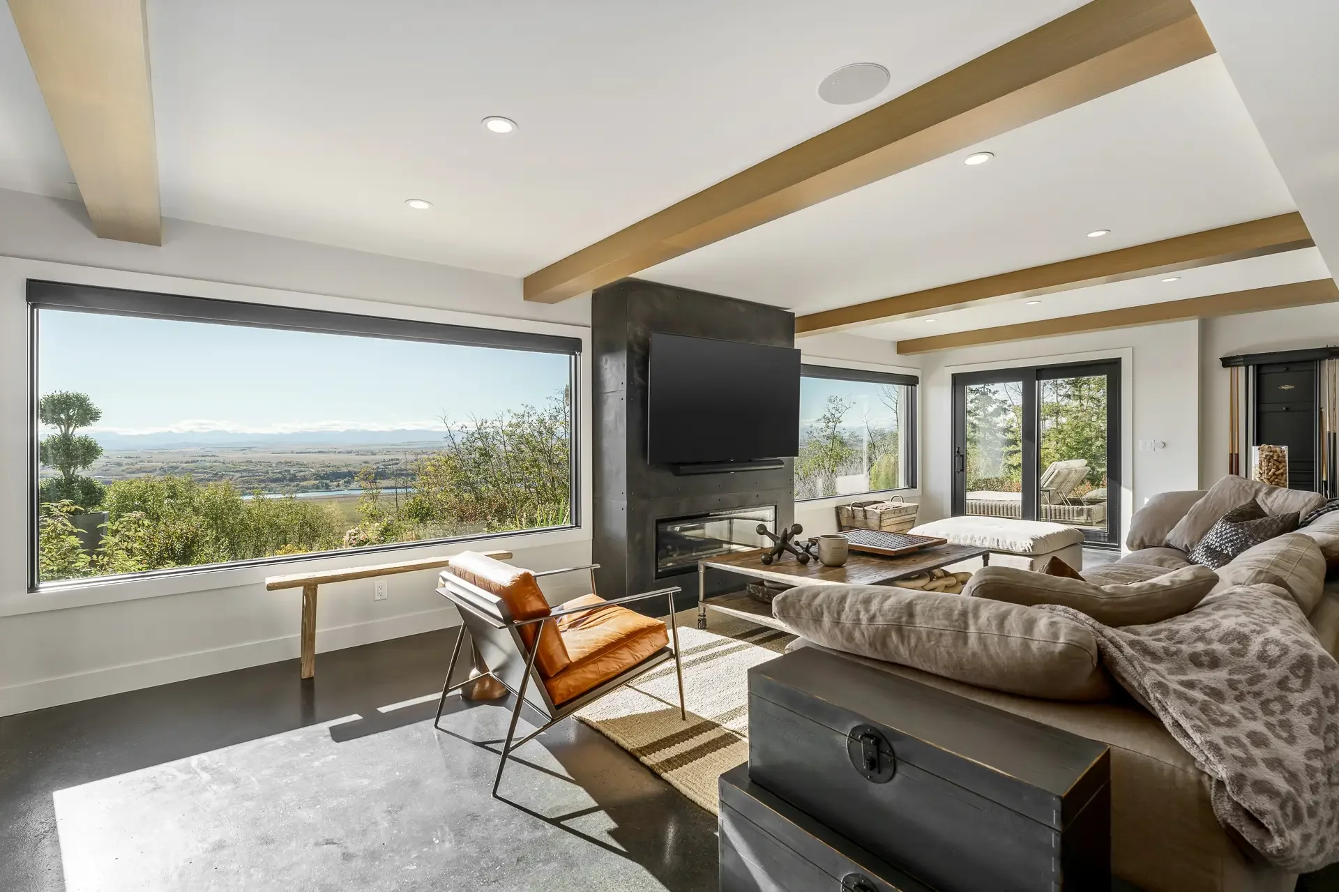 Living room with large windows showing a scenic landscape, featuring a beige sectional sofa, a wooden coffee table, a black fireplace with a mounted flat-screen TV, wooden ceiling beams, and outdoor seating visible through sliding glass doors.