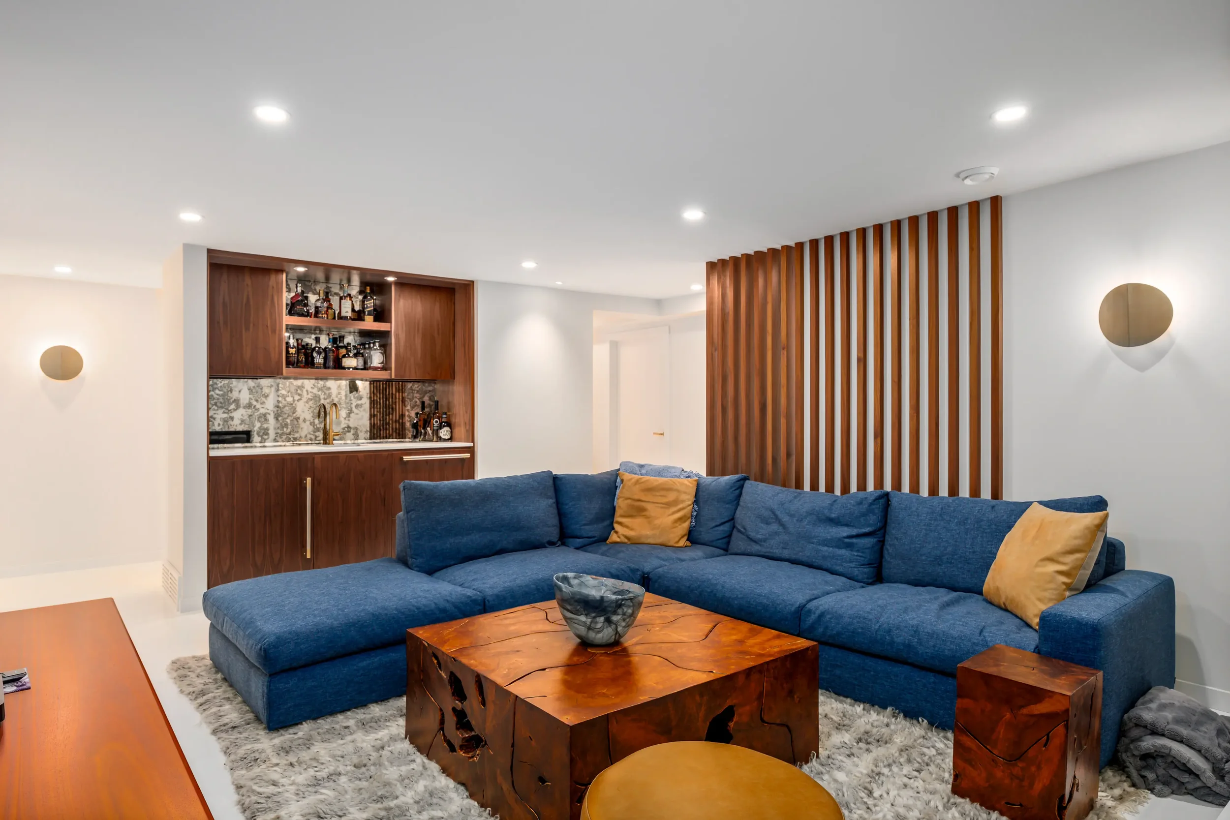 Living room with blue sectional sofa with yellow pillows, wooden coffee table, small side table, and modern wall sconces, with a built-in bar area with liquor bottles and wooden slats dividing the space.