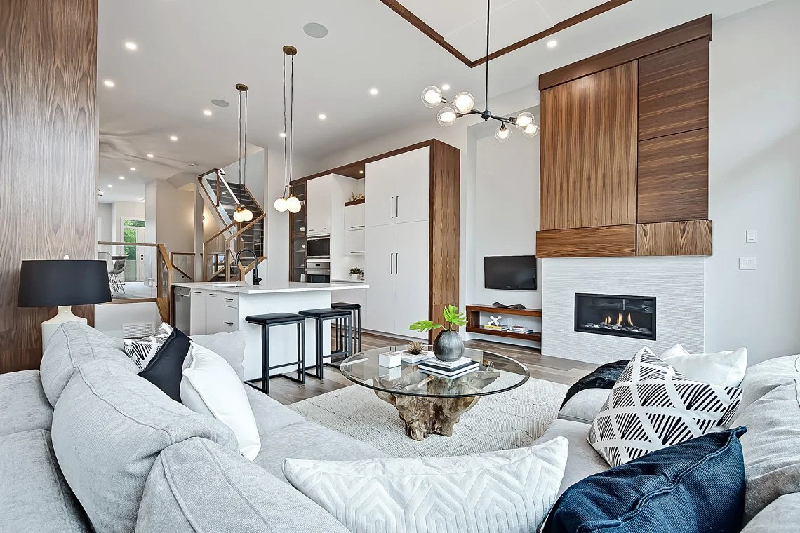 Modern open-concept living room with a fireplace, white walls, a gray sofa with patterned pillows, a glass coffee table with a potted plant, and a white kitchen with wooden accents and pendant lighting.