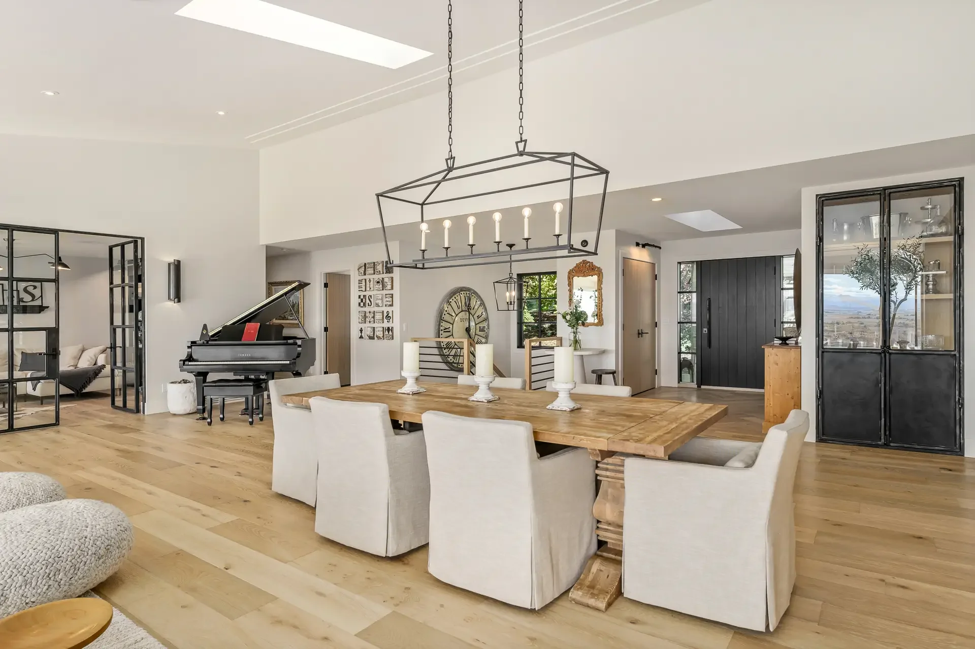 Open-concept living and dining area with a dining table, white chairs, a black grand piano, and glass-front cabinets, featuring modern decor and large windows overlooking the outdoors.