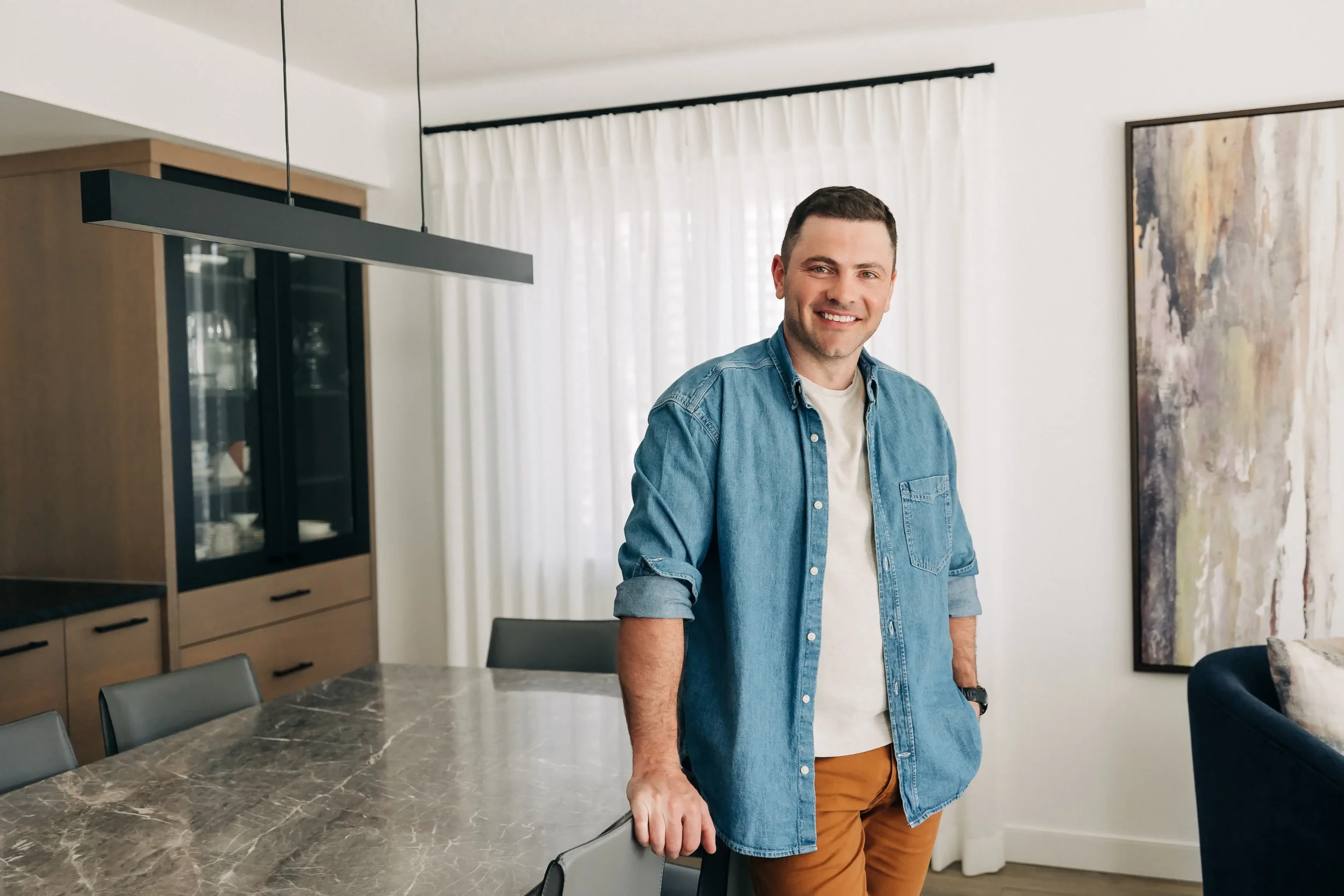 A man with short dark hair and light skin, smiling, standing in a modern dining room with a marble table, chairs, and a large abstract painting on the wall. He is wearing a denim shirt over a white t-shirt and tan pants.