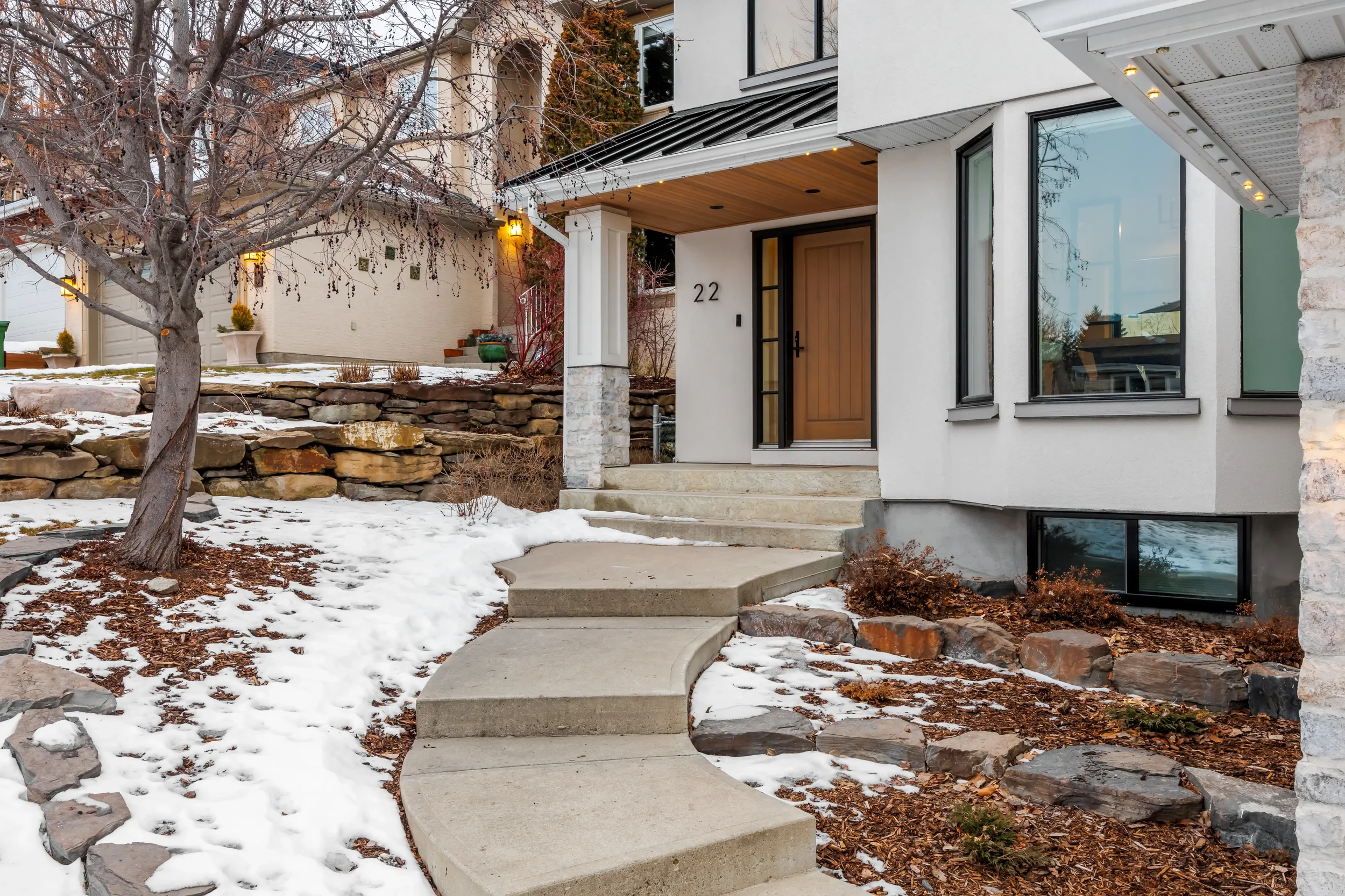 Modern house with large windows, wooden front door, stone steps, landscaped yard with a leafless tree, a fan-shaped stone wall, and snow on the ground.