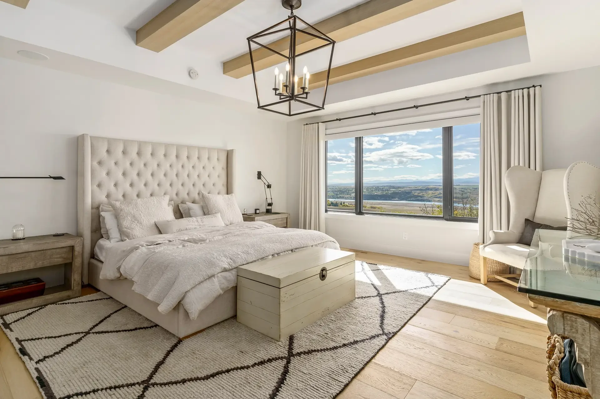 Bright bedroom with beige upholstered bed, white bedding, two nightstands, large window with white curtains showing a scenic view of a river and landscape, beige armchair, ceiling light fixture, and a patterned rug on wooden floor.
