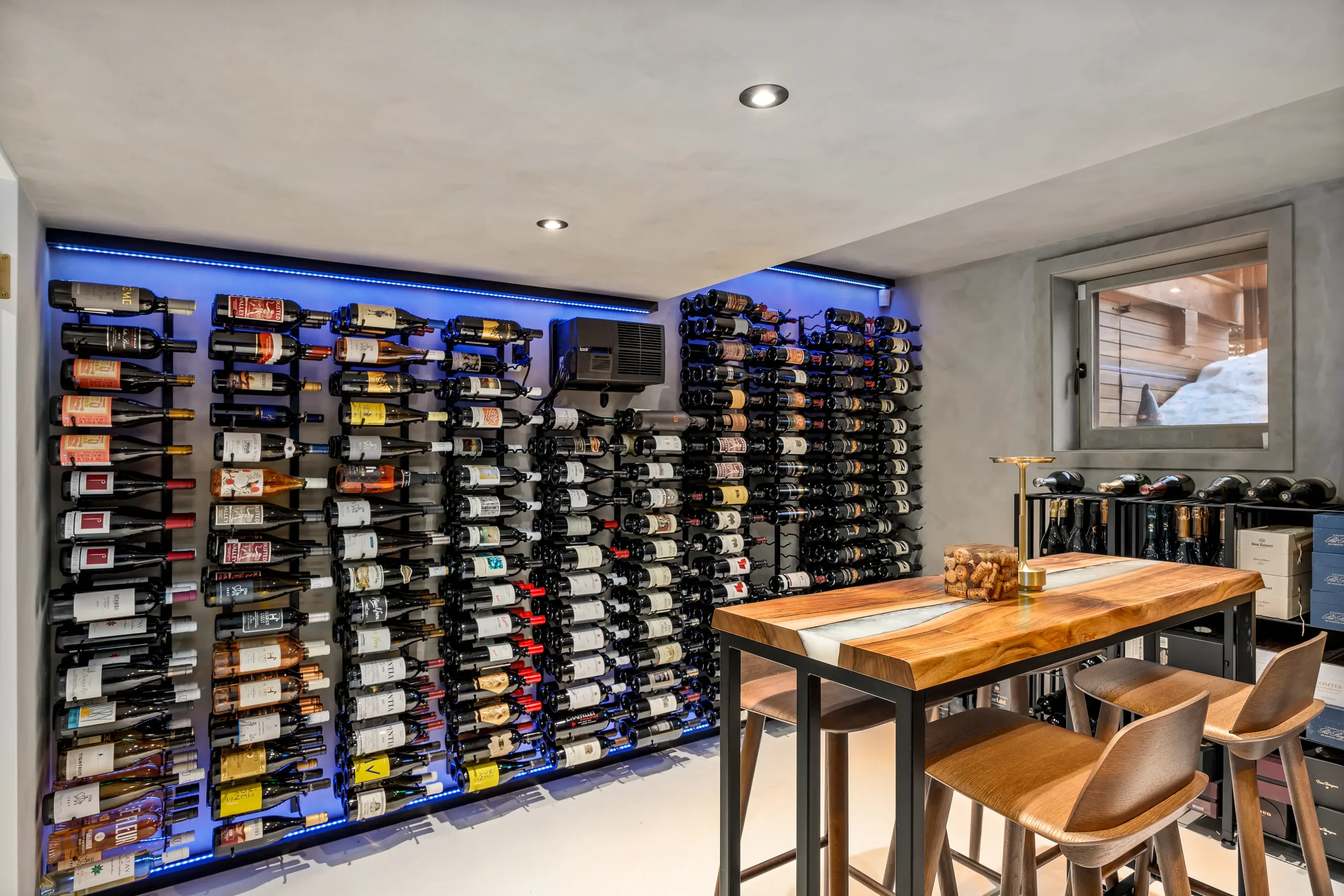 A wine cellar with bottles organized on wall-mounted racks and a wooden table with chairs, a window above the table, and ambient lighting.