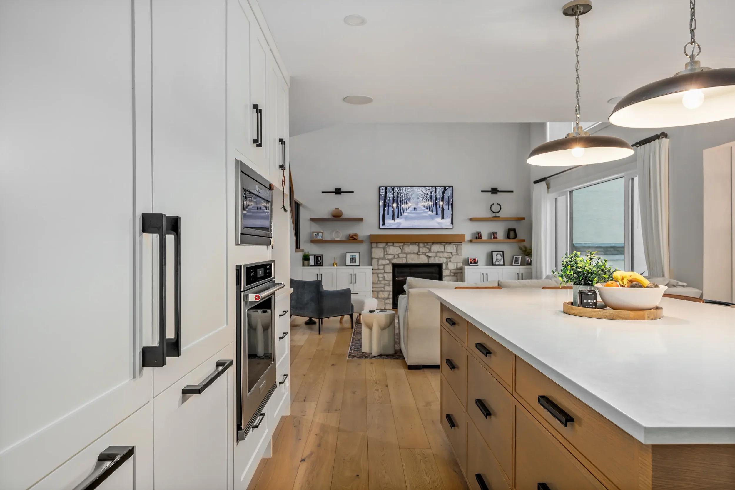 The image shows a modern and bright kitchen with white cabinetry, stainless steel appliances, and a large island with a wooden base. There are two large pendant lights hanging above the island, a bowl of fruit, and a view into a cozy living room with a fireplace, TV, and framed photos. The room has large windows with white curtains and hardwood floors.