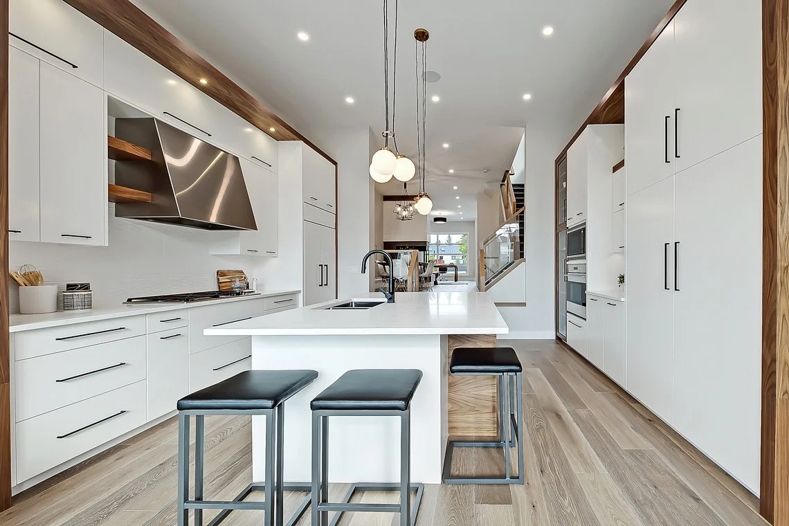 Modern kitchen with white cabinets, black handles, wooden accents, a central island with black stools, and stainless steel appliances. Pendant lights hang over the island, and a view of the dining area and living space is visible in the background.
