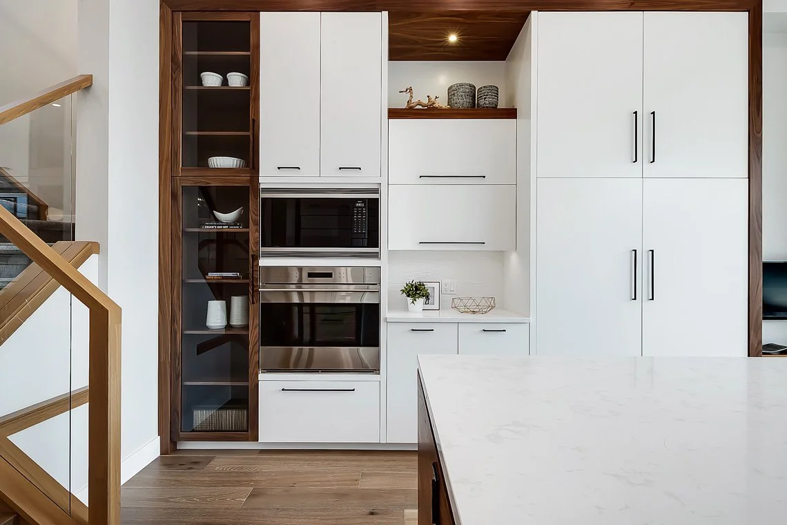 Modern kitchen with white cabinets, built-in stainless steel microwave and oven, open shelving with dishes, decorative items on a shelf, a potted plant on the counter, and a marble kitchen island in the foreground.