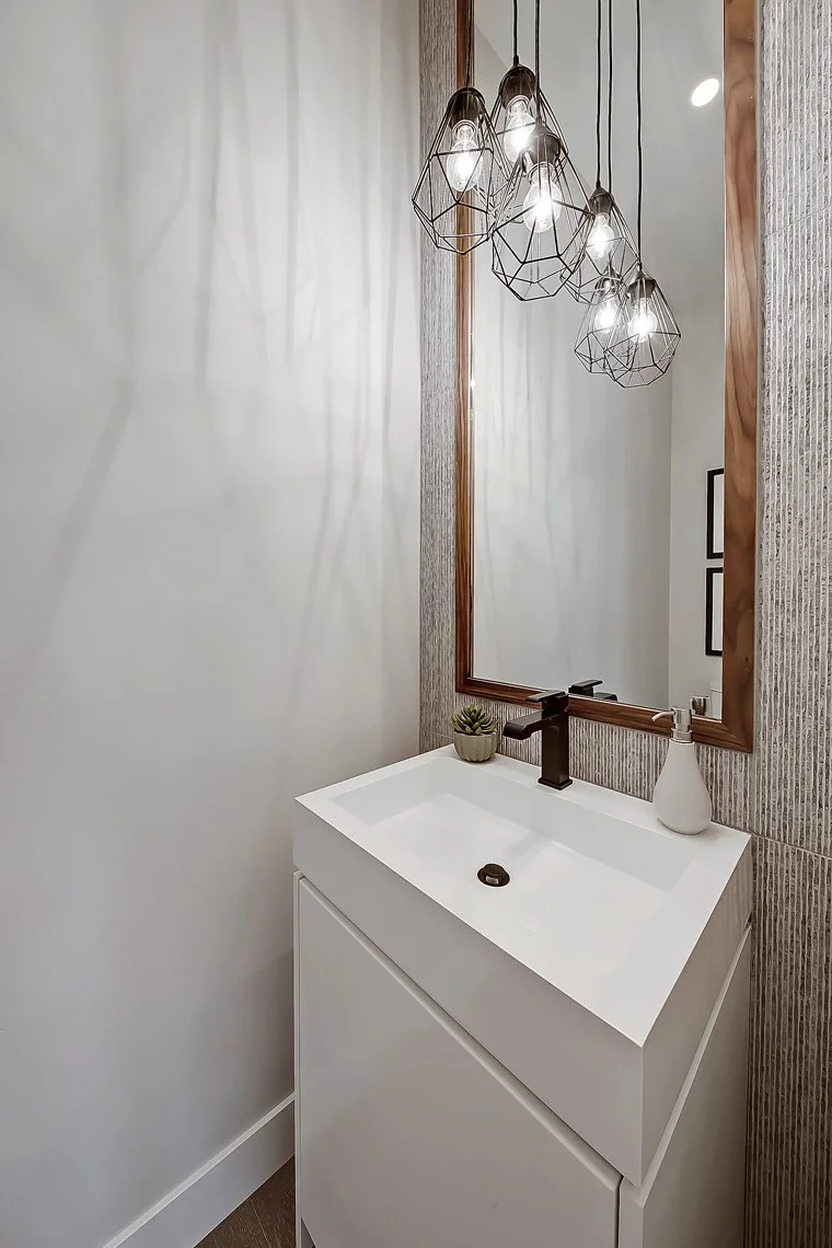 Small white bathroom vanity with a black faucet, a small potted plant, a white soap dispenser, a large mirror with a wooden frame, and a geometric pendant light fixture with exposed bulbs casting shadows on the wall.