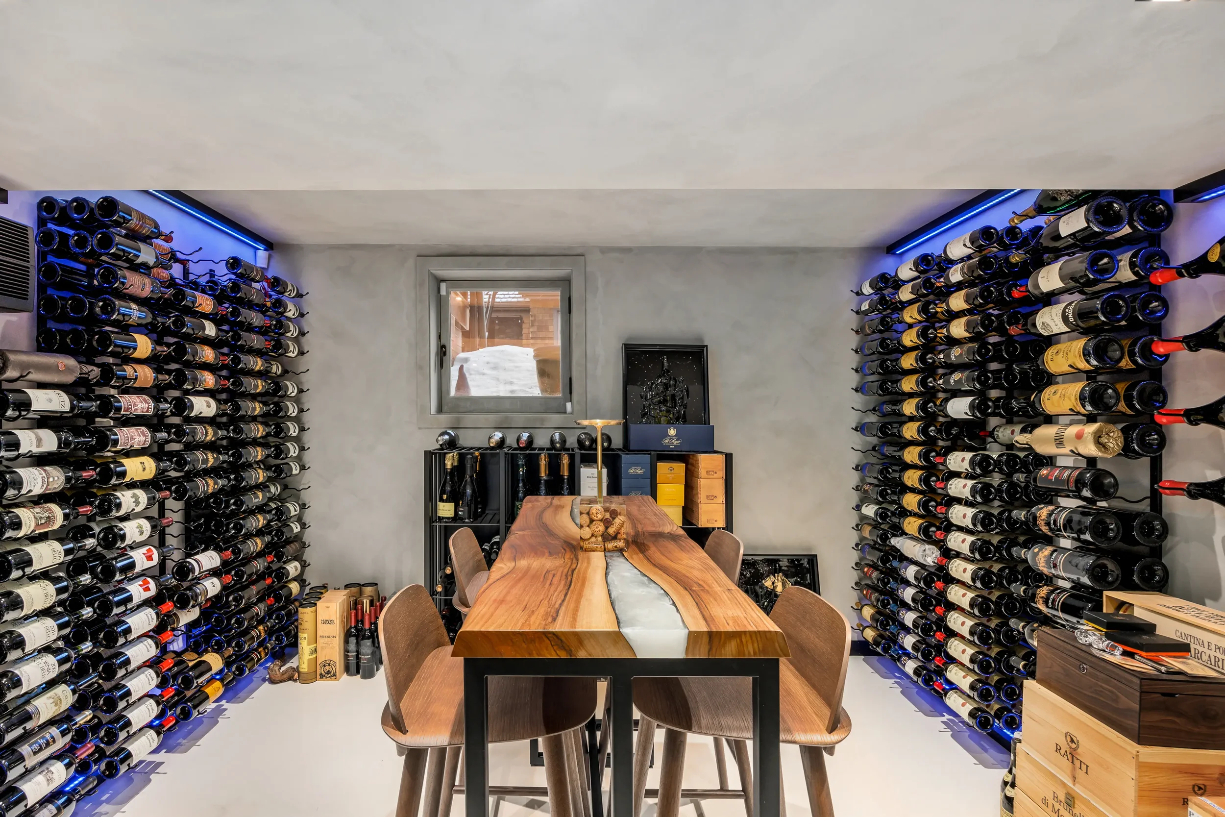 Image of a wine cellar with bottles stored on both sides, a wooden table in the center, surrounded by chairs, a small window, and wine storage boxes on the floor.