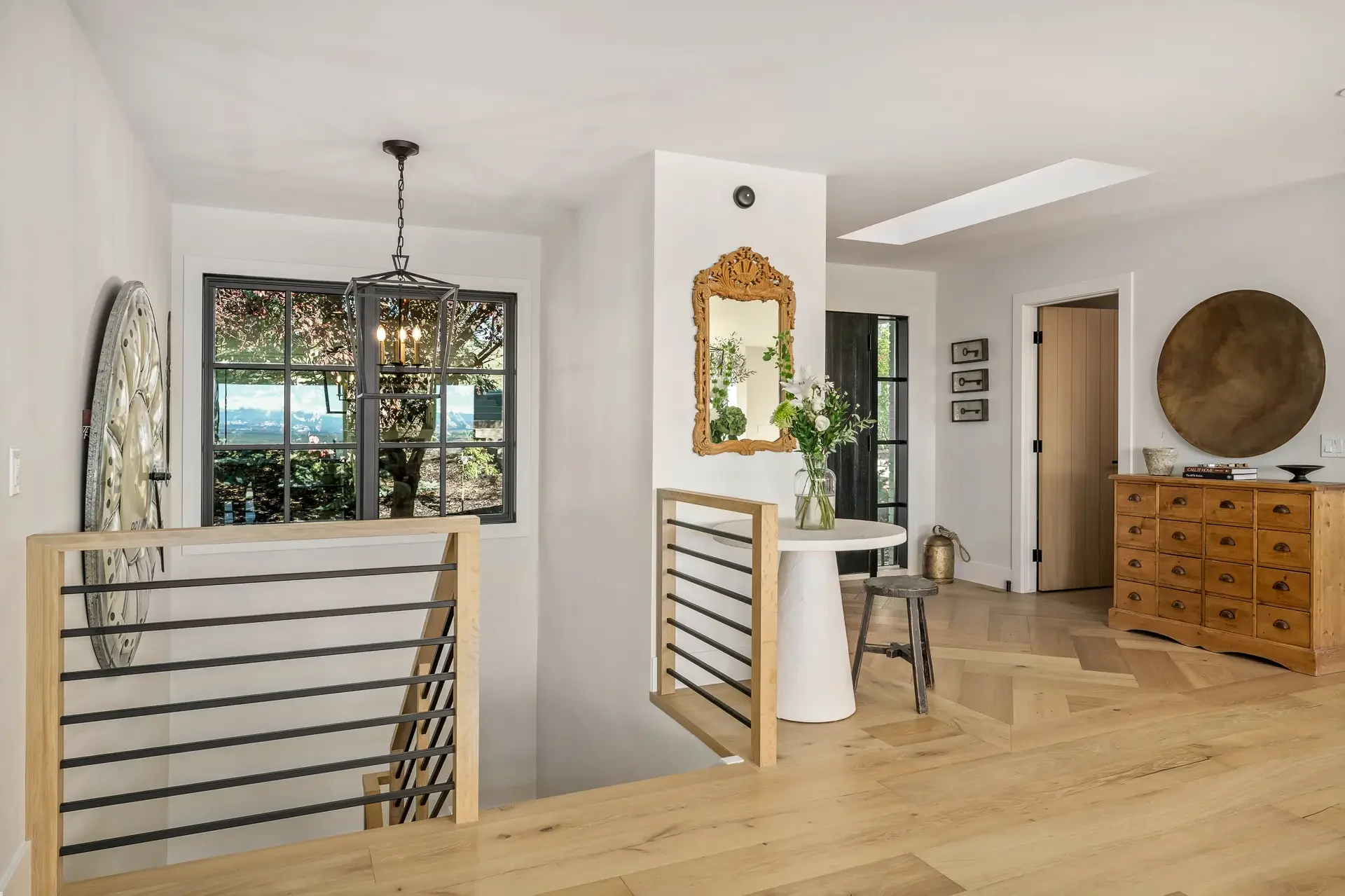 Interior of a modern house with a white wall, wooden furniture, and decorative items. A chandelier hangs in front of the window showing trees and blue sky outside. There's a large ornate mirror with greenery, a wooden chest of drawers with books, bow