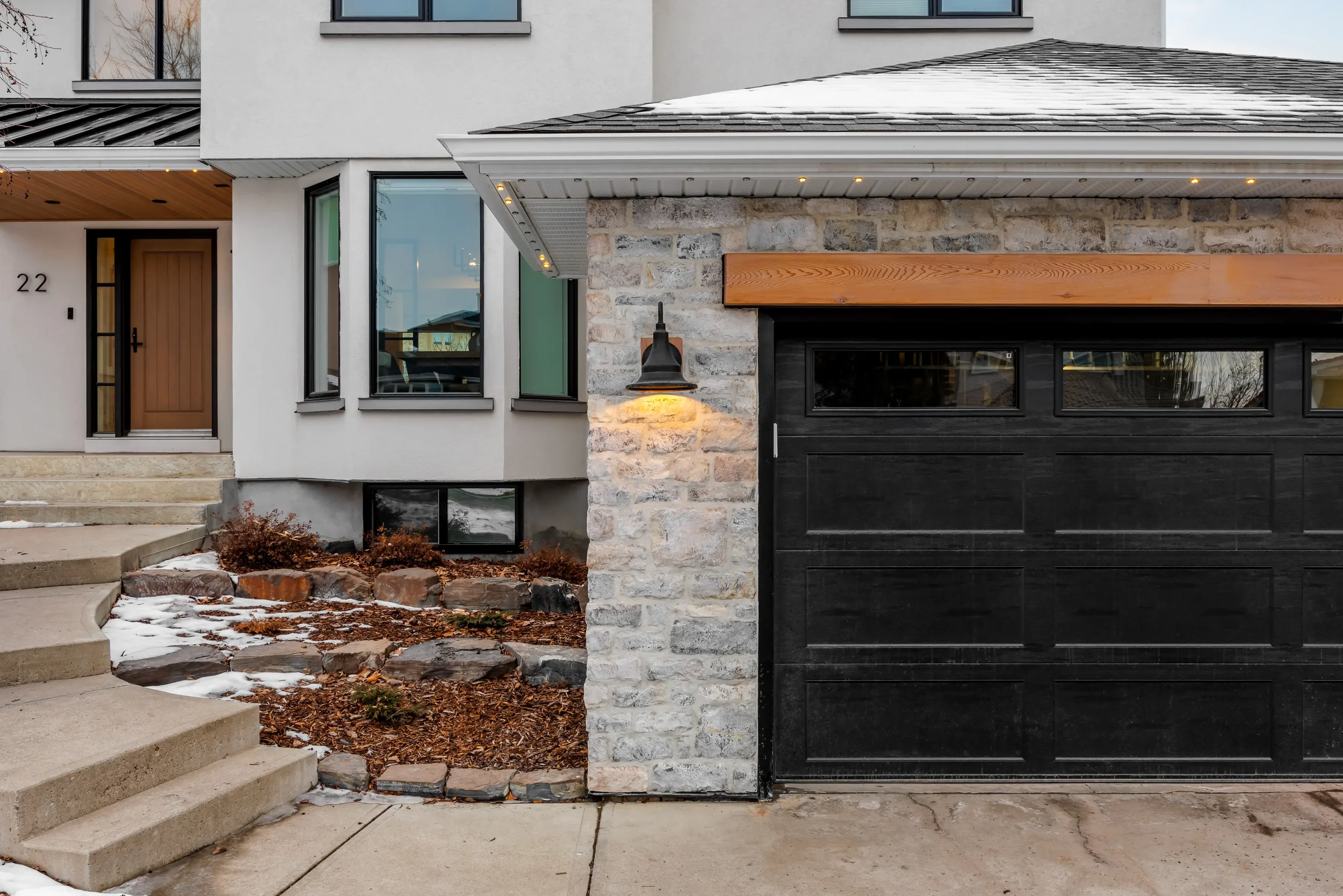 Close-up of a modern house facade with a wooden front door, large windows, a stone and black garage door, and exterior light fixture, with some snow on the ground and steps.