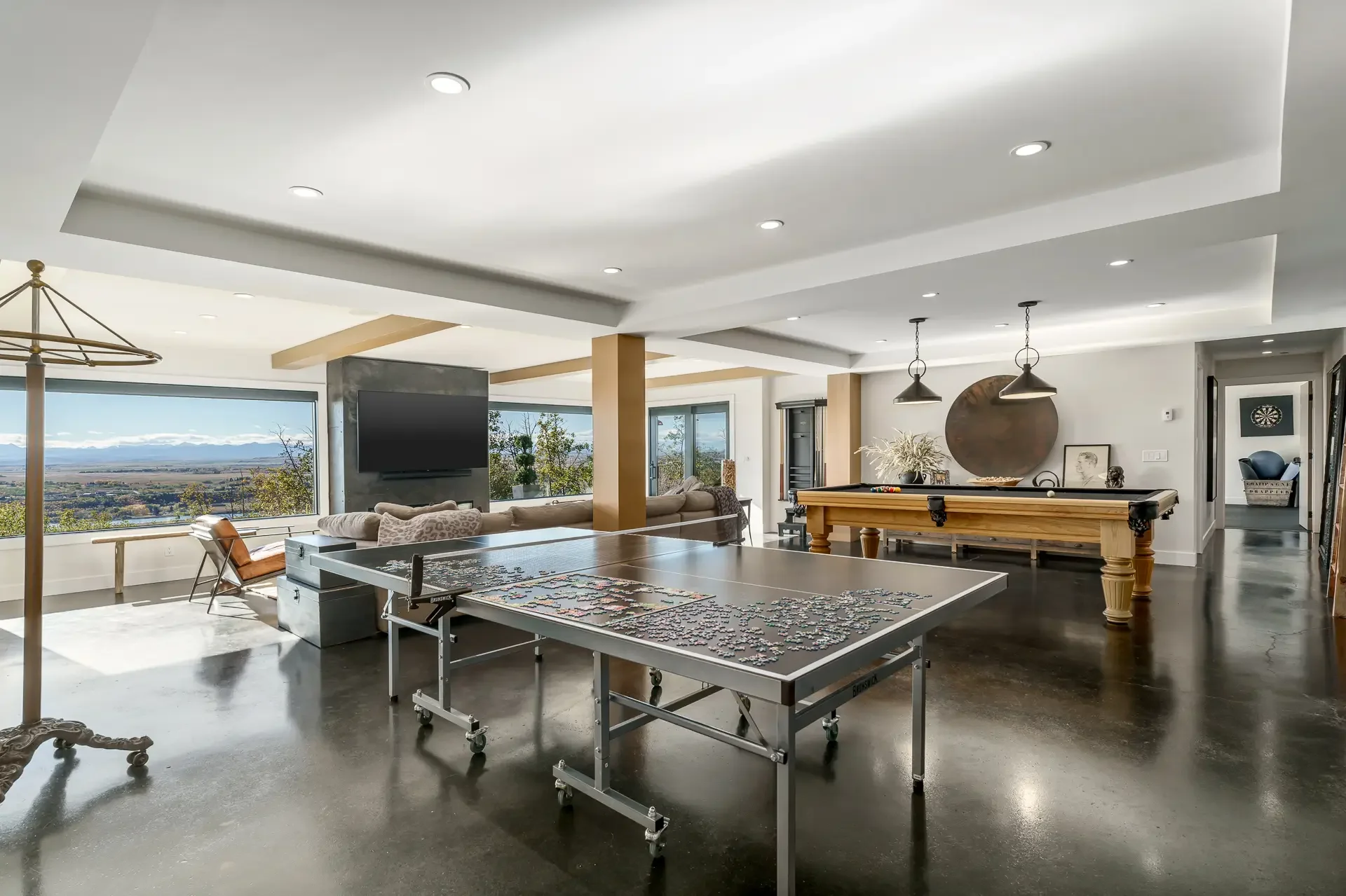 Interior of a modern, spacious living room with large windows, a pool table, a flat-screen TV, and minimalist decor in neutral tones.