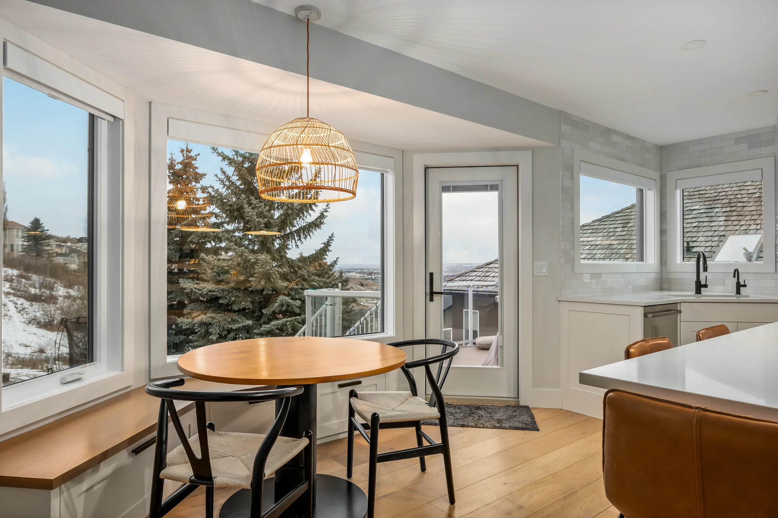 A bright kitchen with large windows showing outdoor trees and rooftops. There is a round wooden table with black chairs, a white countertop with a black faucet, and a wicker hanging light fixture.