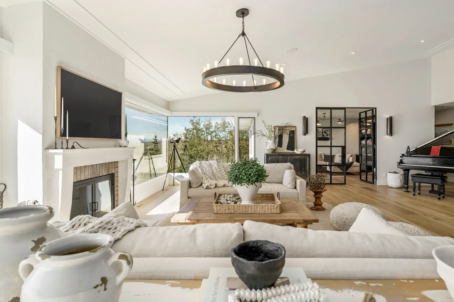 Bright living room with a large window, a chandelier, a television above a fireplace, a black divided door separating spaces, a piano, and various decorative items.