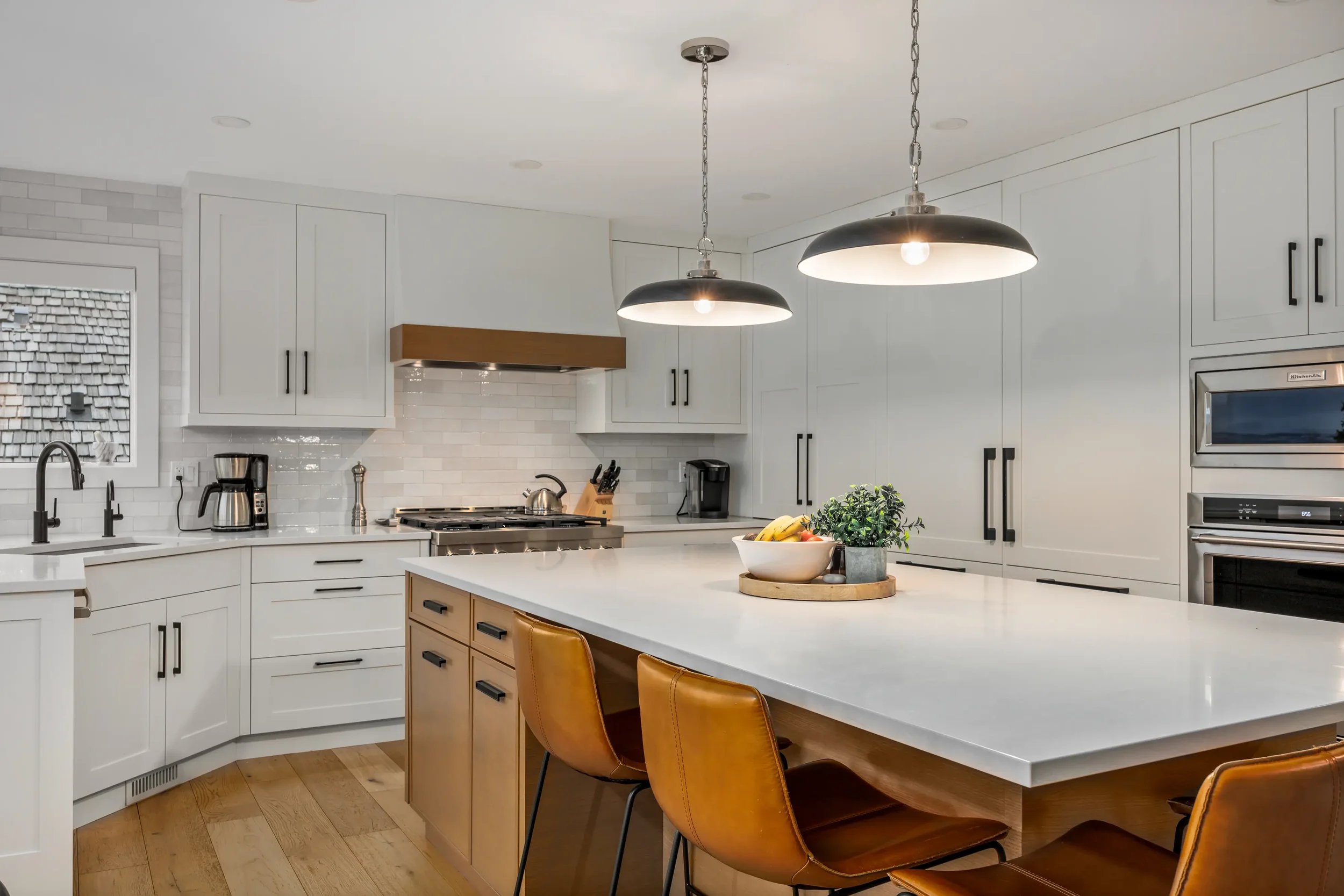 Modern white kitchen with an island, black and gold chairs, pendant lights, stainless steel appliances, and a window with a shingled exterior view.