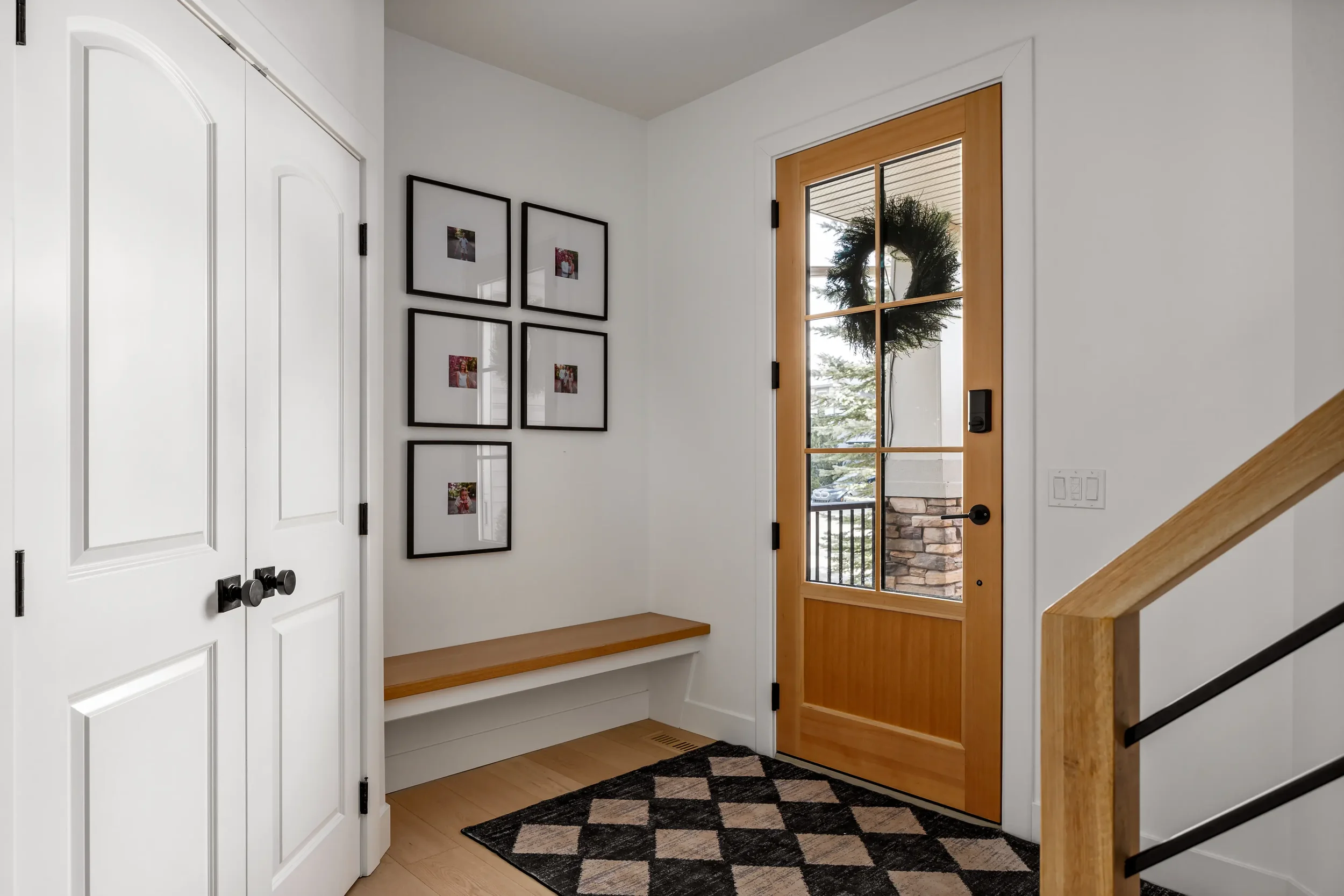 Entryway with a wooden door featuring glass panes, a black handle, and a large holiday wreath hanging outside. Inside, there are white walls with a four-frame photo display, a black and beige patterned rug, a wooden bench underneath the picture displ