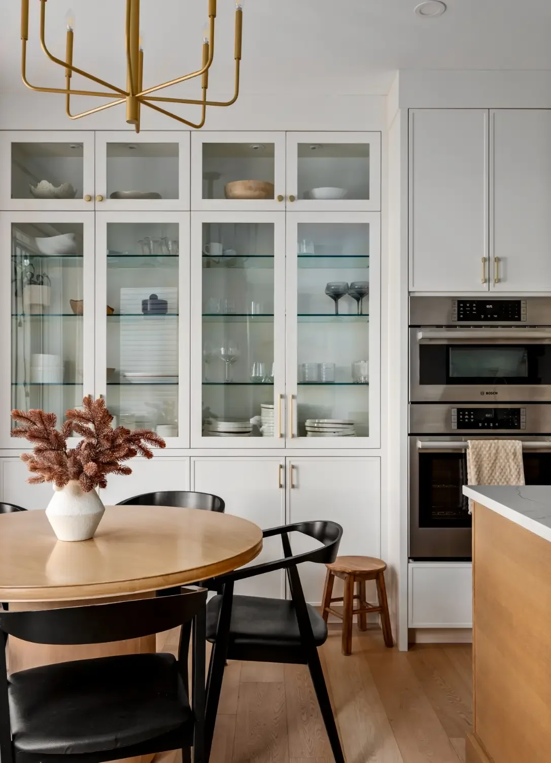 Modern kitchen featuring a white cabinet with glass doors displaying dishes and glassware, stainless steel oven and microwave, wooden dining table with black chairs, and a decorative vase with pink dried flowers.