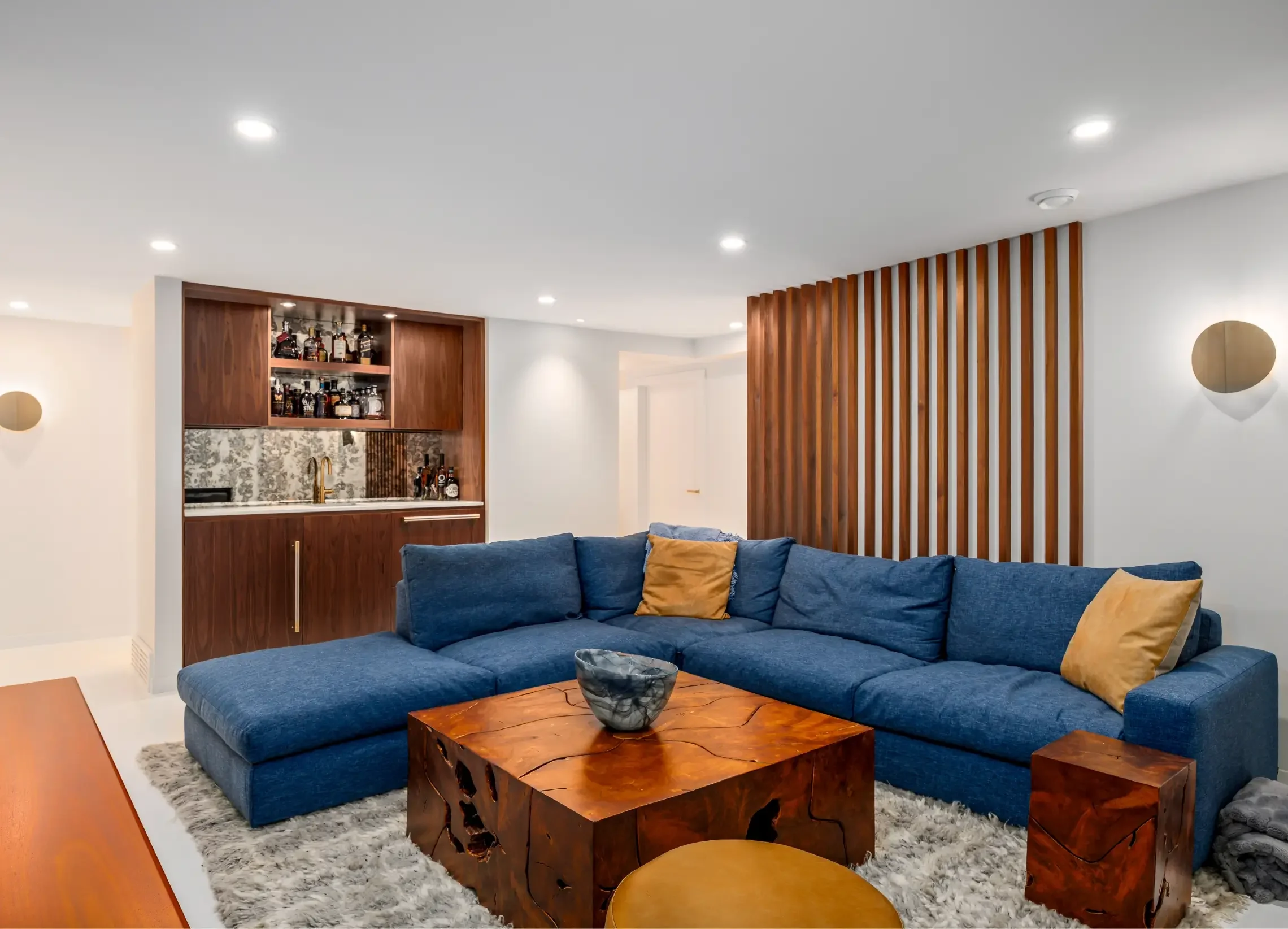 Living room with a blue sectional sofa, a rustic wooden coffee table, and a wooden slat wall divider. There is a built-in bar with bottles and a decorative backsplash in the background. The ceiling has recessed lighting and a white wall with round sconces.