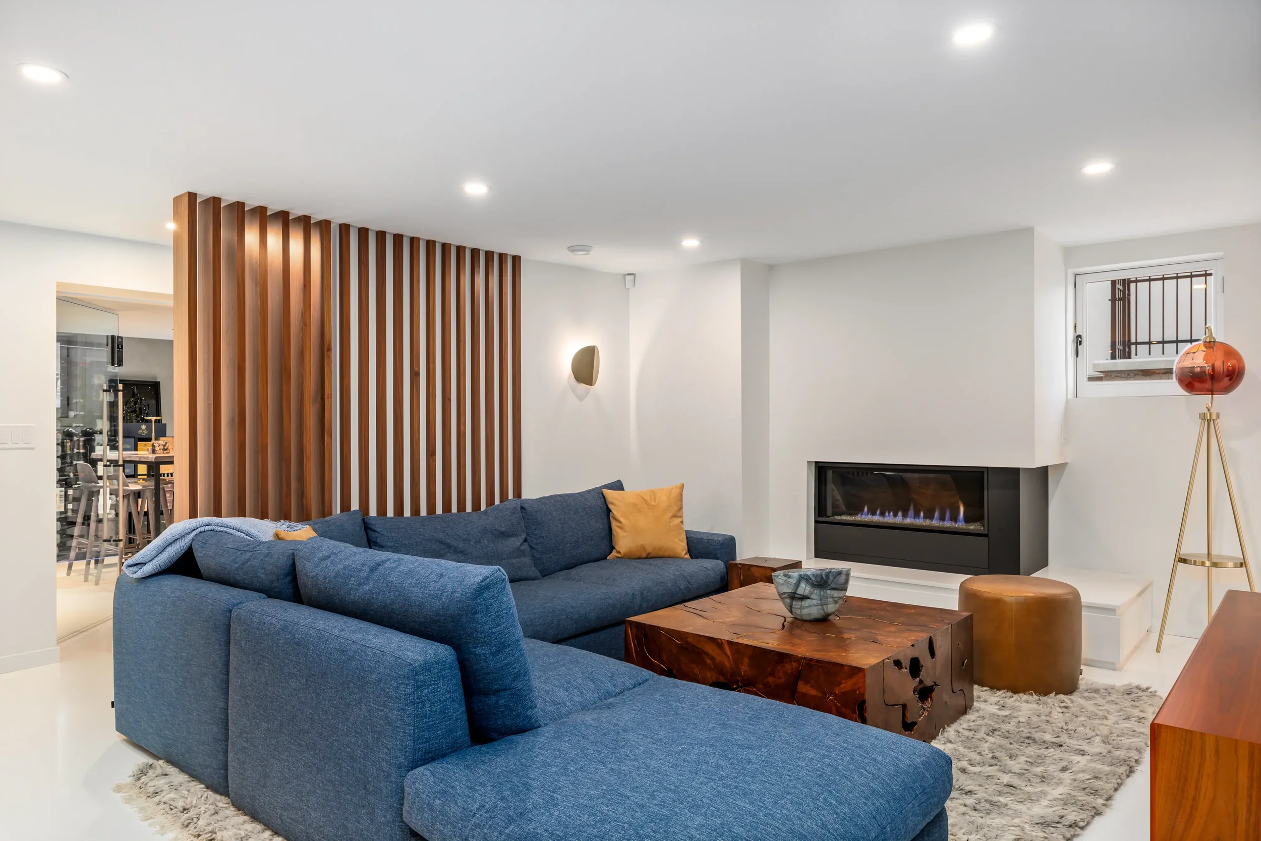 A living room with a blue sectional sofa, wooden coffee table, and modern fireplace. The space has white walls, a window with bars, and a floor lamp with a copper-colored shade. Wooden slats create a partition wall.